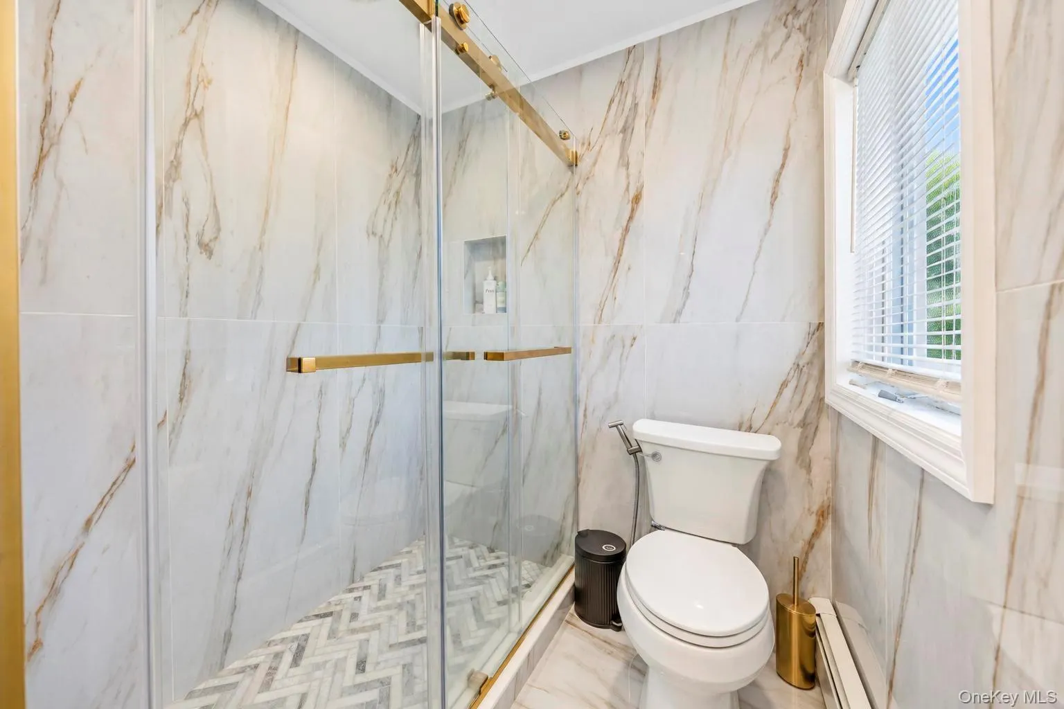 Glass-enclosed shower with gold-toned hardware, marble-look wall tiles, and a herringbone-patterned shower floor Glass-enclosed shower with gold-toned hardware, marble-look wall tiles, and a herringbone-patterned shower floor