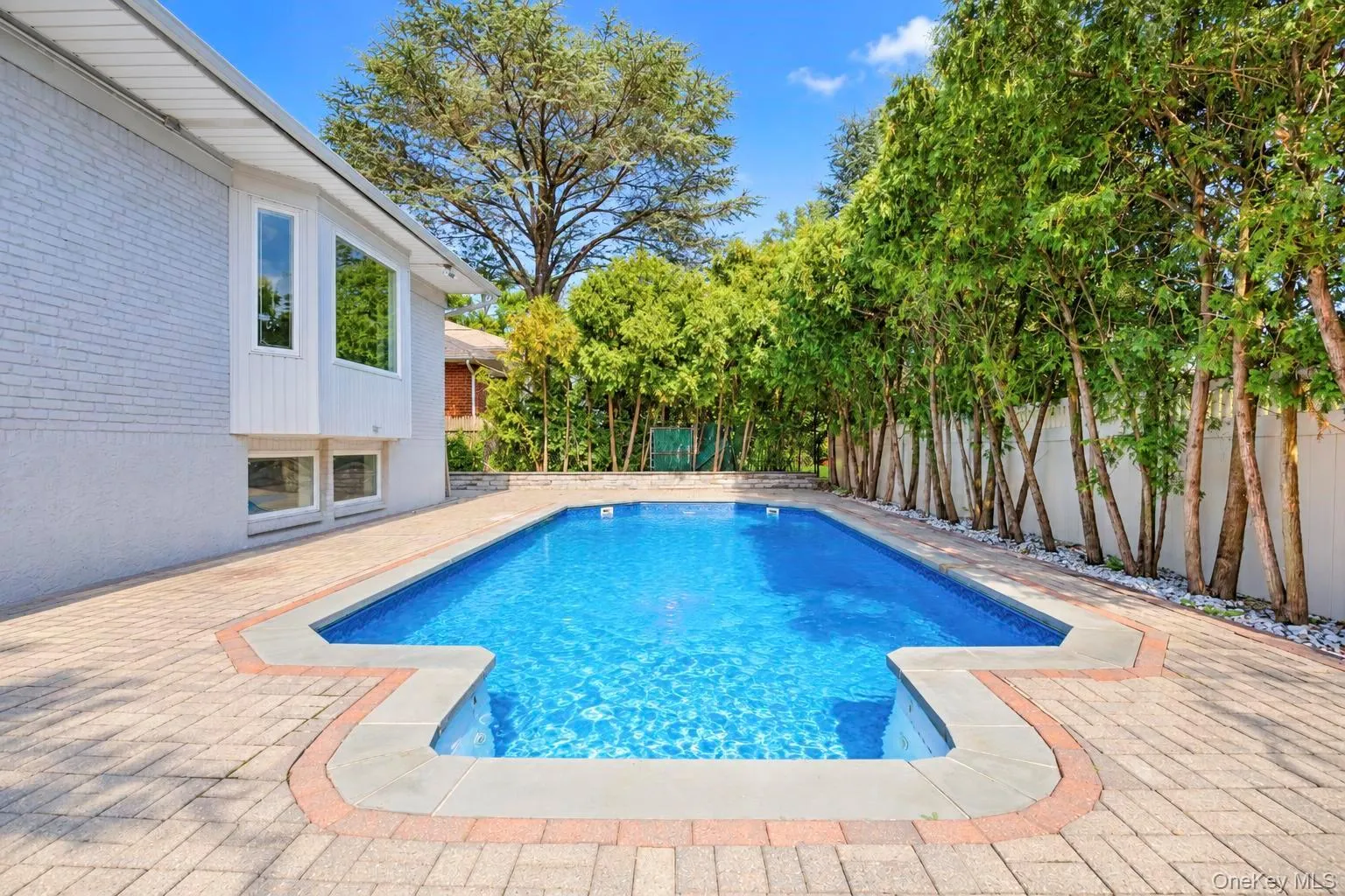 Rectangular swimming pool with integrated corner steps, surrounded by paver patio with contrasting border, mature trees, and a white privacy fence Rectangular swimming pool with integrated corner steps, surrounded by paver patio with contrasting border, mature trees, and a white privacy fence