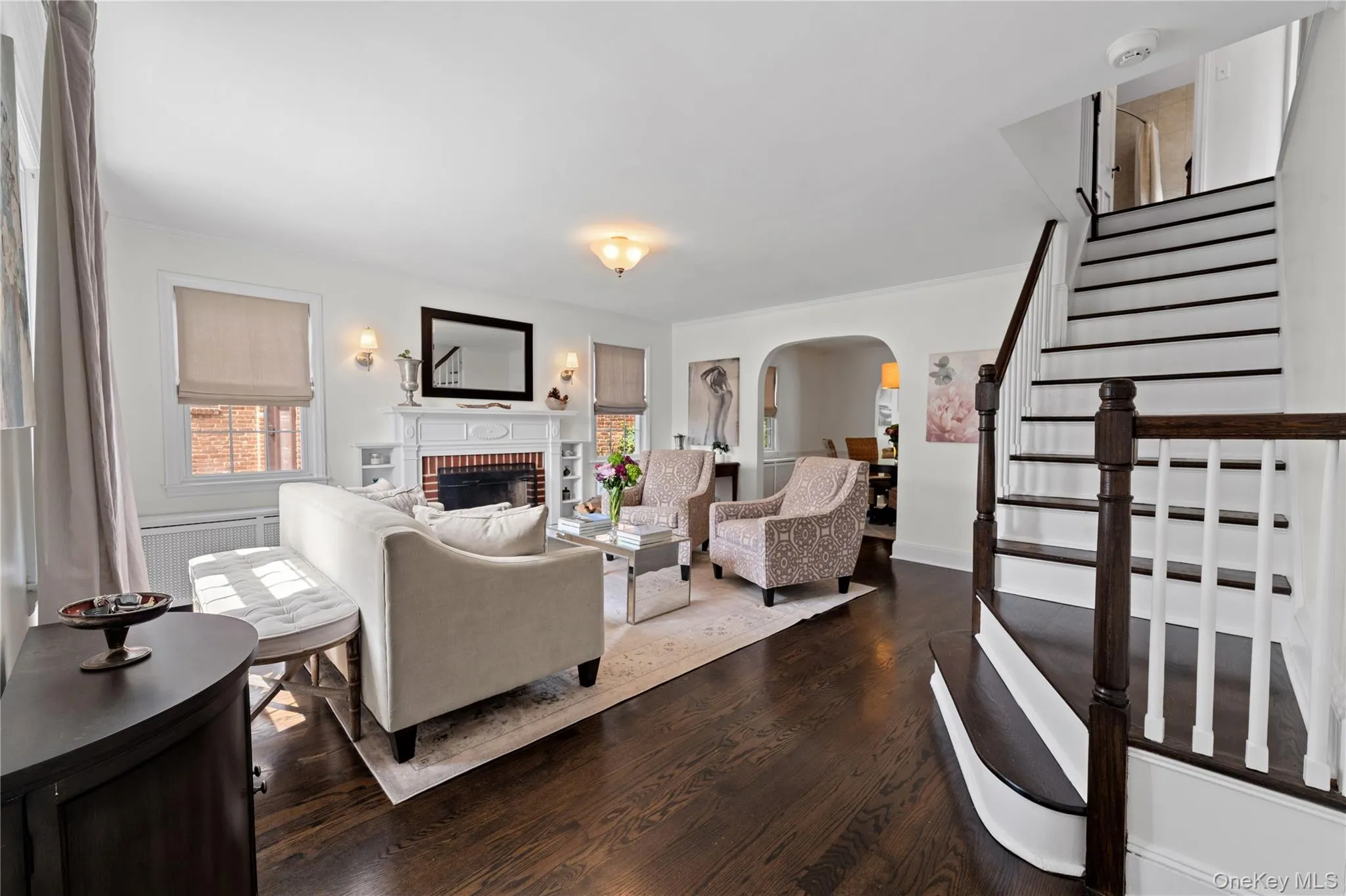 Rich wood-finish flooring throughout, a traditional fireplace with a white mantel, and a gracefully curved staircase with dark wood newel posts and banister Rich wood-finish flooring throughout, a traditional fireplace with a white mantel, and a gracefully curved staircase with dark wood newel posts and banister