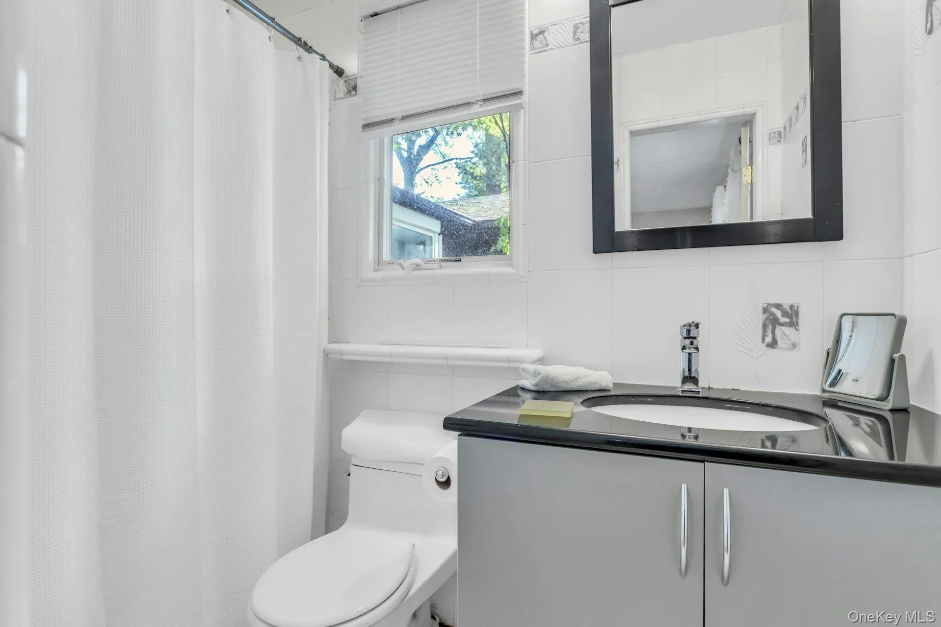 Vanity with a dark countertop and integrated sink, chrome faucet, wall-mounted mirror with a black frame, and a window with blinds Vanity with a dark countertop and integrated sink, chrome faucet, wall-mounted mirror with a black frame, and a window with blinds