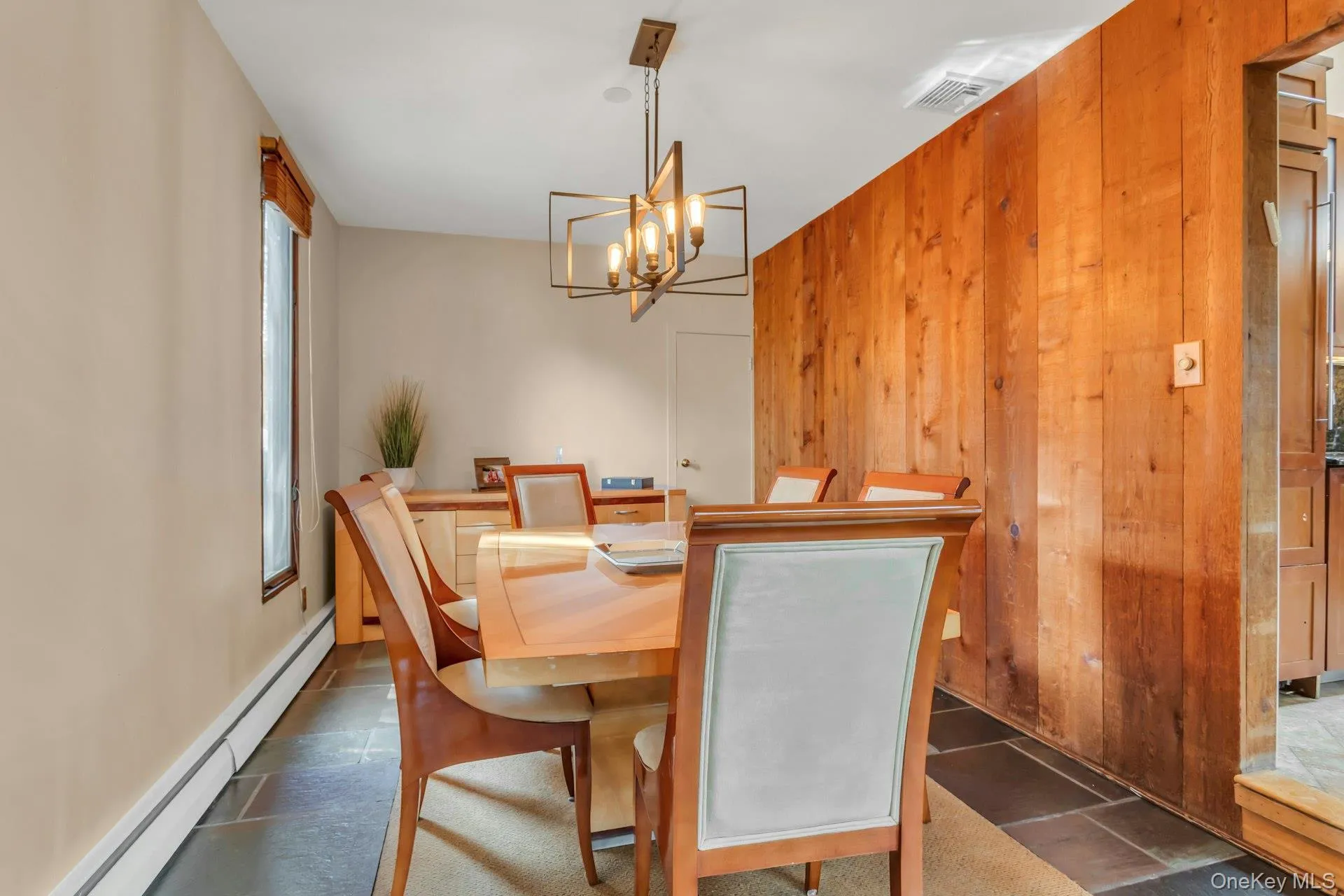 Elegant dining room showcasing warm wood-finish wall paneling, a round wood dining table with upholstered seating, geometric brass pendant lighting fixture, and slate-finish flooring Elegant dining room showcasing warm wood-finish wall paneling, a round wood dining table with upholstered seating, geometric brass pendant lighting fixture, and slate-finish flooring