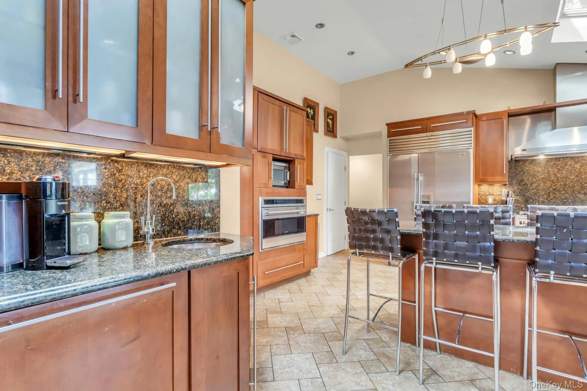 Kitchen featuring granite countertops, integrated stainless steel appliances, wood cabinetry with frosted glass inserts, and tiled flooring Kitchen featuring granite countertops, integrated stainless steel appliances, wood cabinetry with frosted glass inserts, and tiled flooring