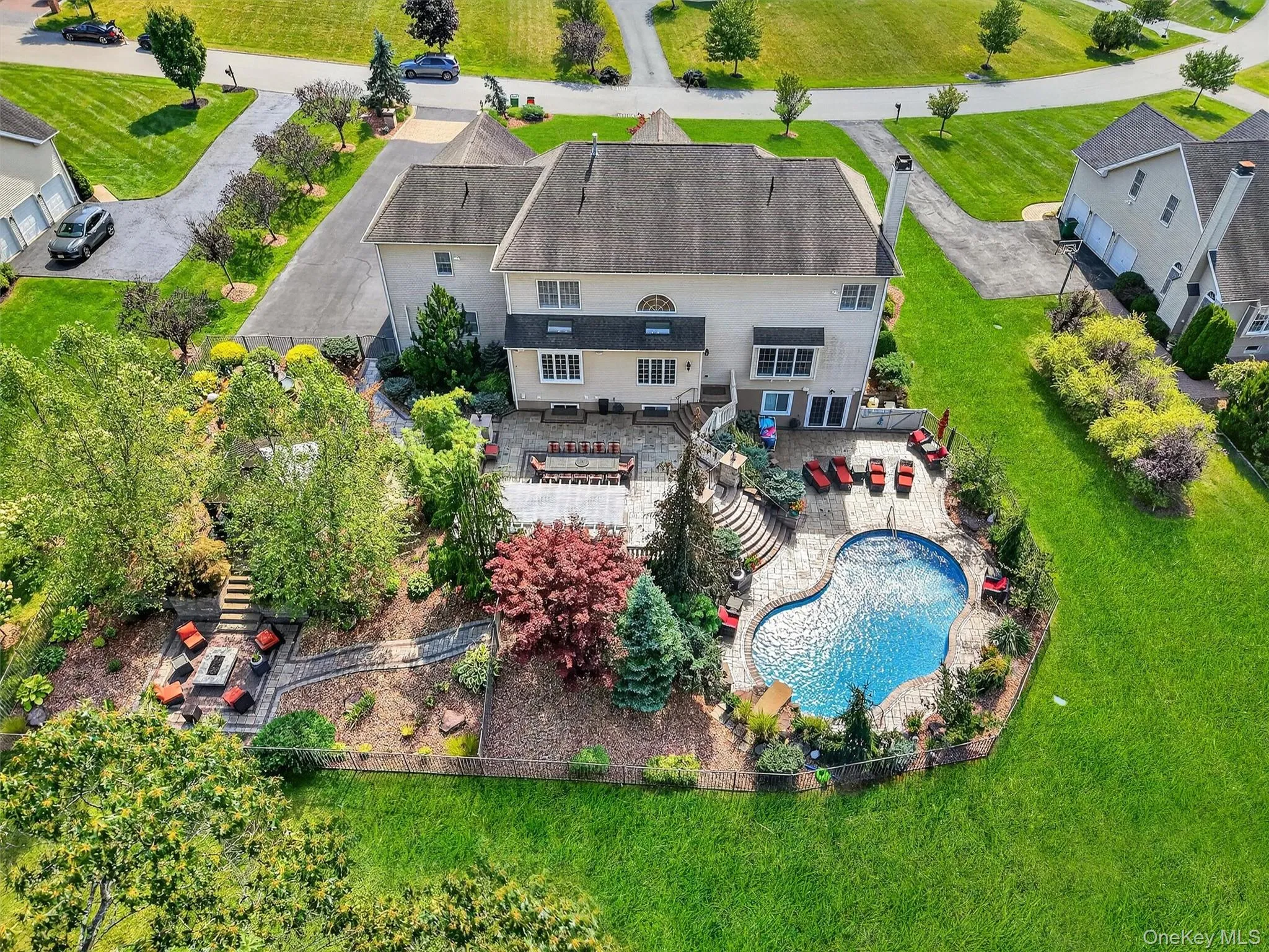 This elevated view highlights the grandeur of a meticulously landscaped backyard that blends privacy with upscale outdoor living. At the heart of the space lies a beautifully contoured in-ground pool surrounded by an expansive paver patio, where plush red lounge seating and umbrella-shaded areas create multiple zones for relaxation and sun-soaked leisure. The multi-tiered layout flows gracefully from the home’s rear entrance down to the pool area, featuring custom stonework, ornamental landscaping, and winding garden paths. A fully furnished dining area and pergola-covered sitting space offer an exquisite setting for alfresco dining or evening gatherings under the stars, while the backyard itself is enclosed with tasteful fencing and lush plantings, ensuring both seclusion and charm. To the left, a private firepit lounge nestled among mature trees offers a cozy hideaway, perfect for quiet nights or intimate conversations, all surrounded by nature’s beauty. This elevated view highlights the grandeur of a meticulously landscaped backyard that blends privacy with upscale outdoor living. At the heart of the space lies a beautifully contoured in-ground pool surrounded by an expansive paver patio, where plush red lounge seating and umbrella-shaded areas create multiple zones for relaxation and sun-soaked leisure. The multi-tiered layout flows gracefully from the home’s rear entrance down to the pool area, featuring custom stonework, ornamental landscaping, and winding garden paths. A fully furnished dining area and pergola-covered sitting space offer an exquisite setting for alfresco dining or evening gatherings under the stars, while the backyard itself is enclosed with tasteful fencing and lush plantings, ensuring both seclusion and charm. To the left, a private firepit lounge nestled among mature trees offers a cozy hideaway, perfect for quiet nights or intimate conversations, all surrounded by nature’s beauty.