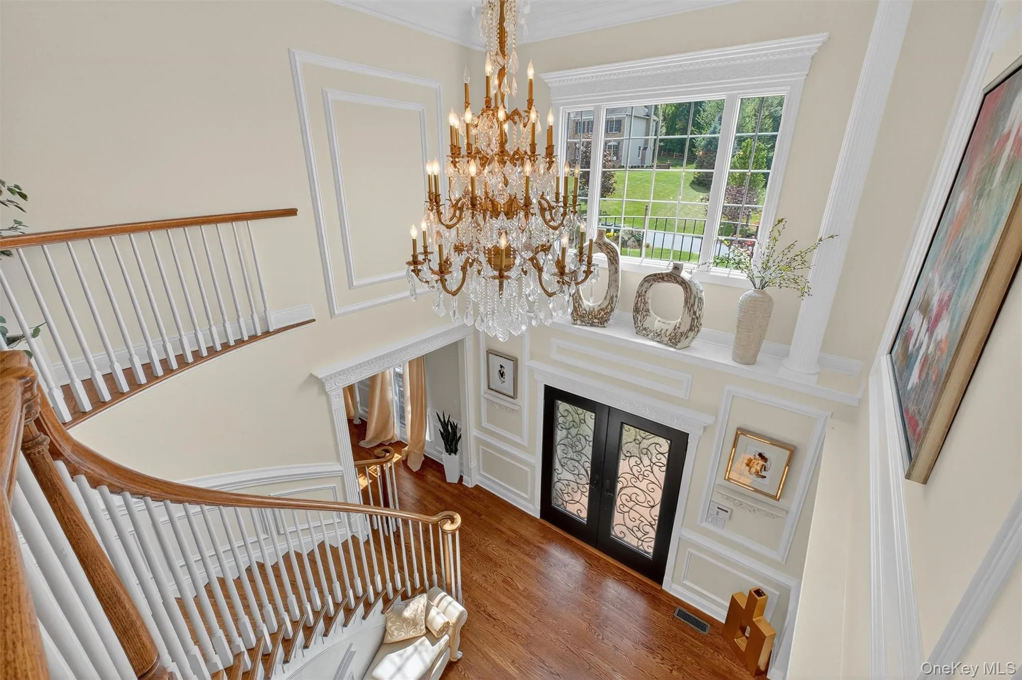 This sweeping staircase is a true statement piece, seamlessly connecting the first and second floors with elegance and grandeur. A brilliant oversized chandelier, operated by a panel lift for easy care, cascades light over the space, creating a regal ambiance fit for a queen. The spectacular triple window unit with decorative accent ledge floods the landing with natural light, highlighting the wood paneling, shadowbox molding, crown detailing, and rich hardwood floors that continue throughout the second level. The oversized landing, just outside the master suite, offers a sense of drama and sophistication—making this staircase not only functional but a breathtaking focal point of the home. This sweeping staircase is a true statement piece, seamlessly connecting the first and second floors with elegance and grandeur. A brilliant oversized chandelier, operated by a panel lift for easy care, cascades light over the space, creating a regal ambiance fit for a queen. The spectacular triple window unit with decorative accent ledge floods the landing with natural light, highlighting the wood paneling, shadowbox molding, crown detailing, and rich hardwood floors that continue throughout the second level. The oversized landing, just outside the master suite, offers a sense of drama and sophistication—making this staircase not only functional but a breathtaking focal point of the home.