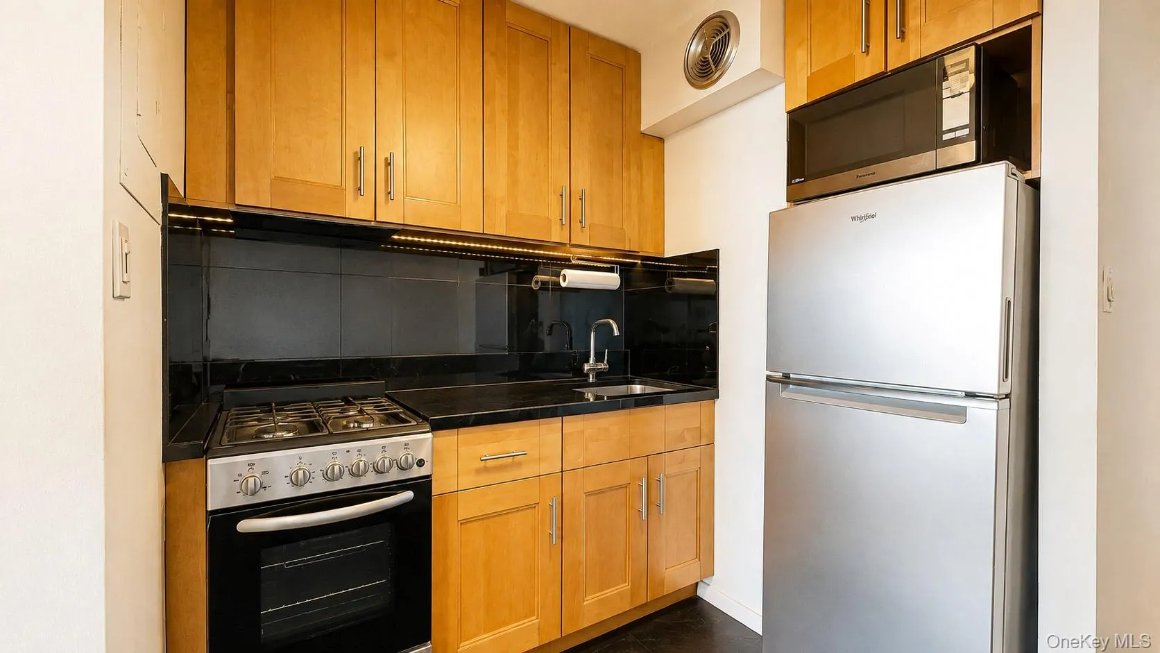 Kitchenette featuring wood-finish cabinetry, sleek Black Granite countertops, a stainless steel refrigerator, and a gas range Kitchenette featuring wood-finish cabinetry, sleek Black Granite countertops, a stainless steel refrigerator, and a gas range