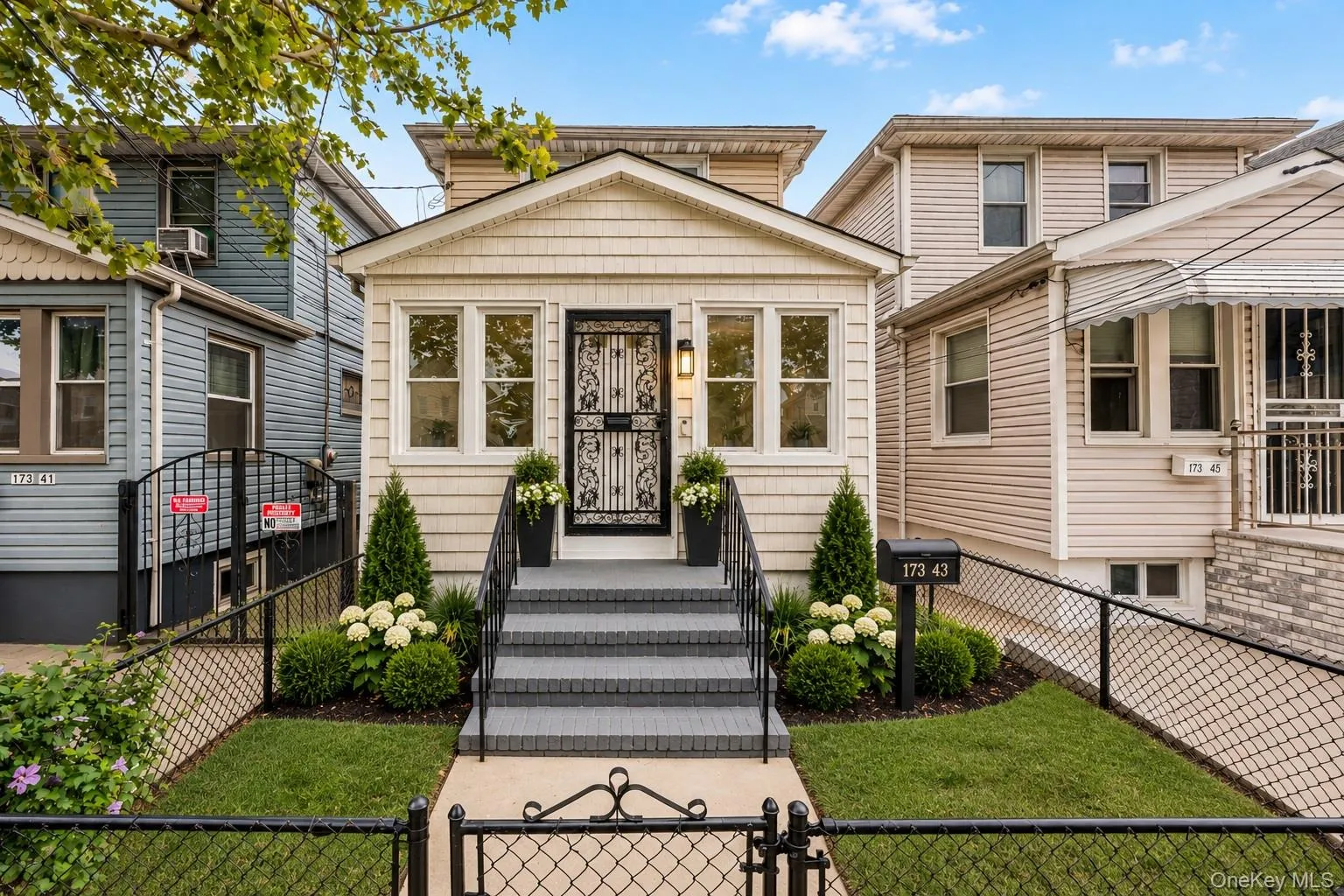 Paved entryway leading to a staircase with black railings, a black security door, and a manicured front yard with mature plantings and a wrought iron fence Paved entryway leading to a staircase with black railings, a black security door, and a manicured front yard with mature plantings and a wrought iron fence