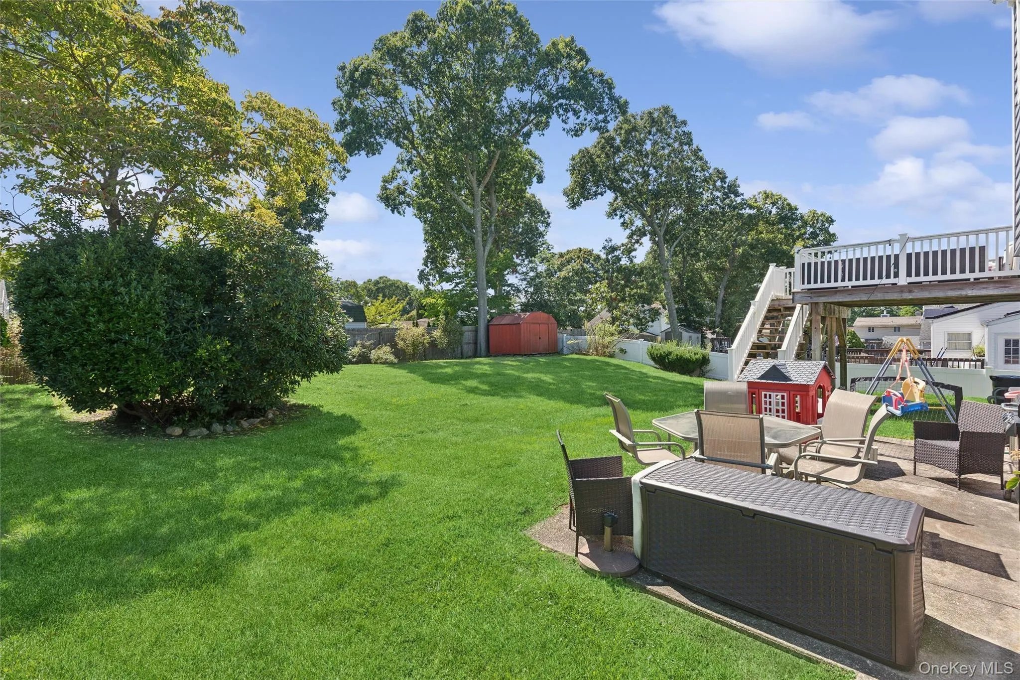 Expansive green lawn with mature trees, featuring a concrete patio, elevated deck with white railing, and exterior staircase Expansive green lawn with mature trees, featuring a concrete patio, elevated deck with white railing, and exterior staircase