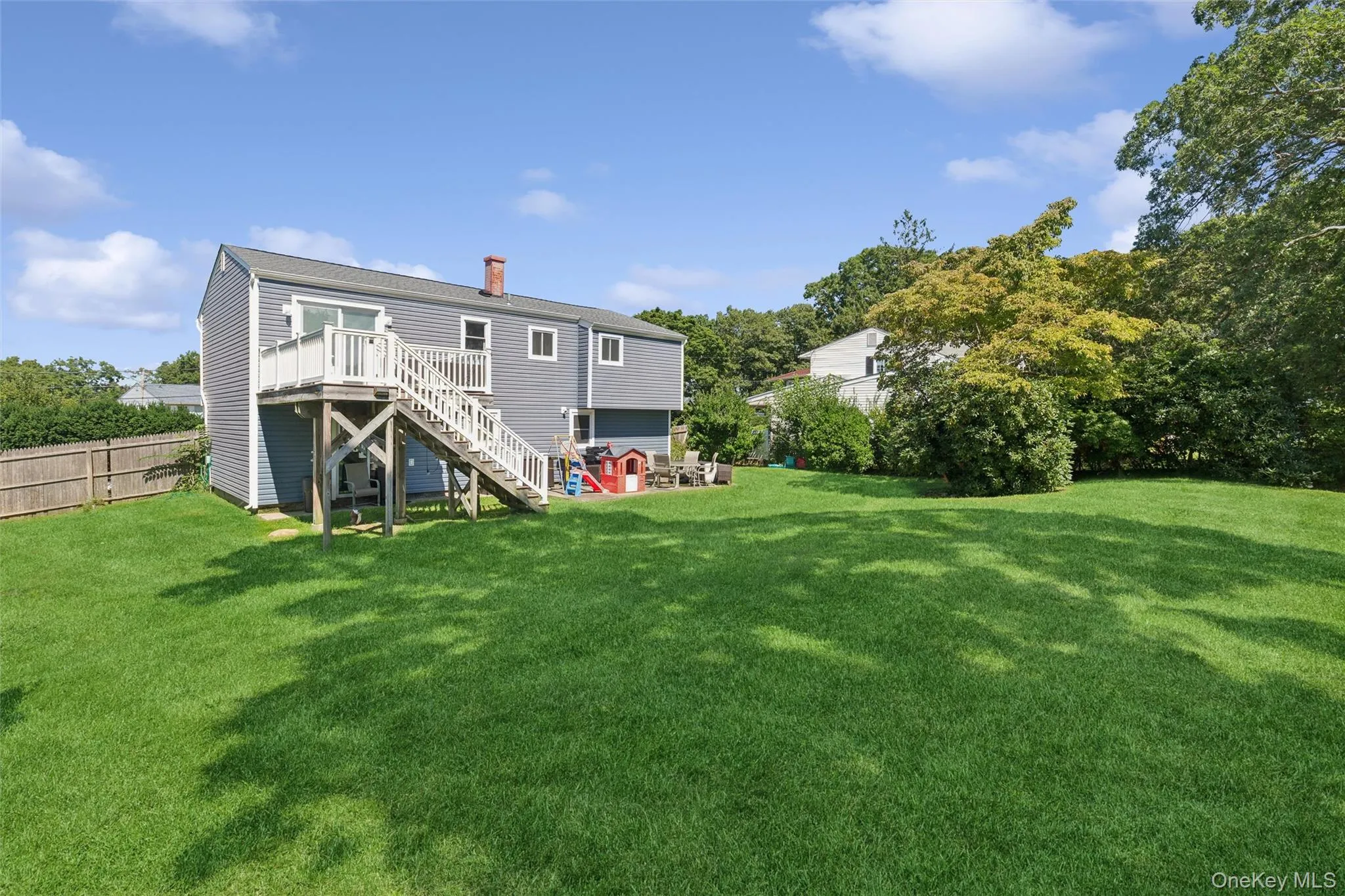 Expansive rear lawn with mature perimeter trees, featuring a raised deck with staircase, and blue siding Expansive rear lawn with mature perimeter trees, featuring a raised deck with staircase, and blue siding