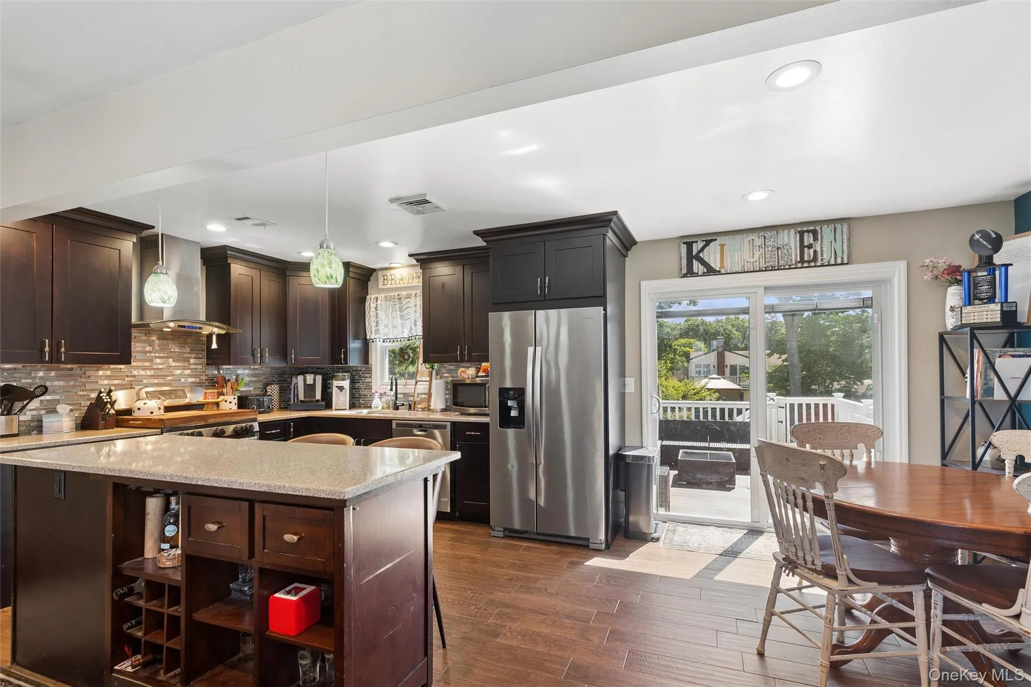 Kitchen featuring dark wood cabinetry, a central island with a light-toned countertop, stainless steel appliances, and wood-finish flooring Kitchen featuring dark wood cabinetry, a central island with a light-toned countertop, stainless steel appliances, and wood-finish flooring