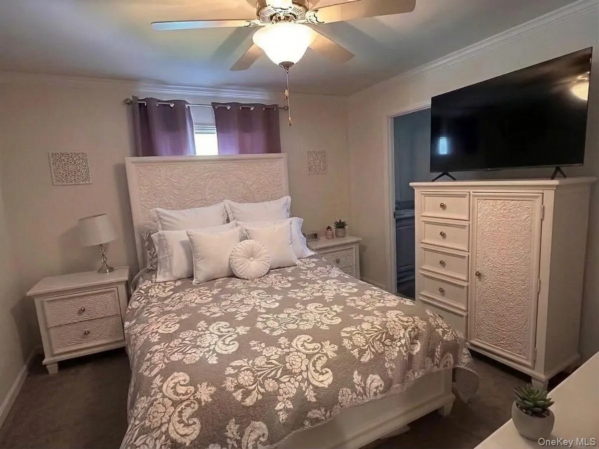 Cream-colored walls with crown molding, ceiling fan with integrated lighting, and a detailed bed frame. Cream-colored walls with crown molding, ceiling fan with integrated lighting, and a detailed bed frame.