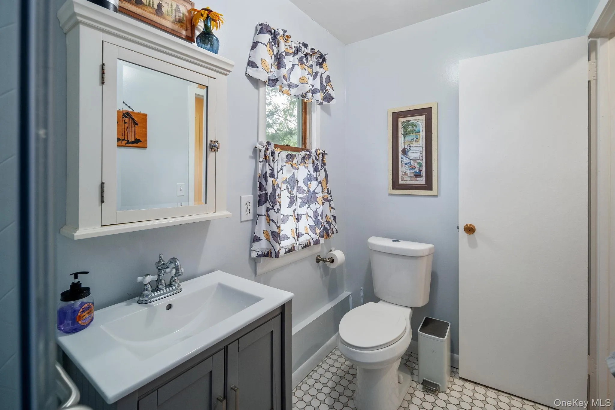 White hexagon tile flooring, a gray vanity with a white integrated sink, a mirrored medicine cabinet, a window, and a white toilet White hexagon tile flooring, a gray vanity with a white integrated sink, a mirrored medicine cabinet, a window, and a white toilet
