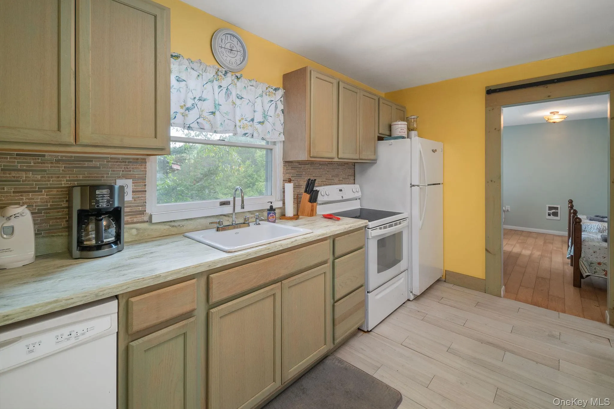 Kitchen featuring light wood-finish cabinetry, a textured tile backsplash, a white porcelain sink, and light-toned countertops Kitchen featuring light wood-finish cabinetry, a textured tile backsplash, a white porcelain sink, and light-toned countertops