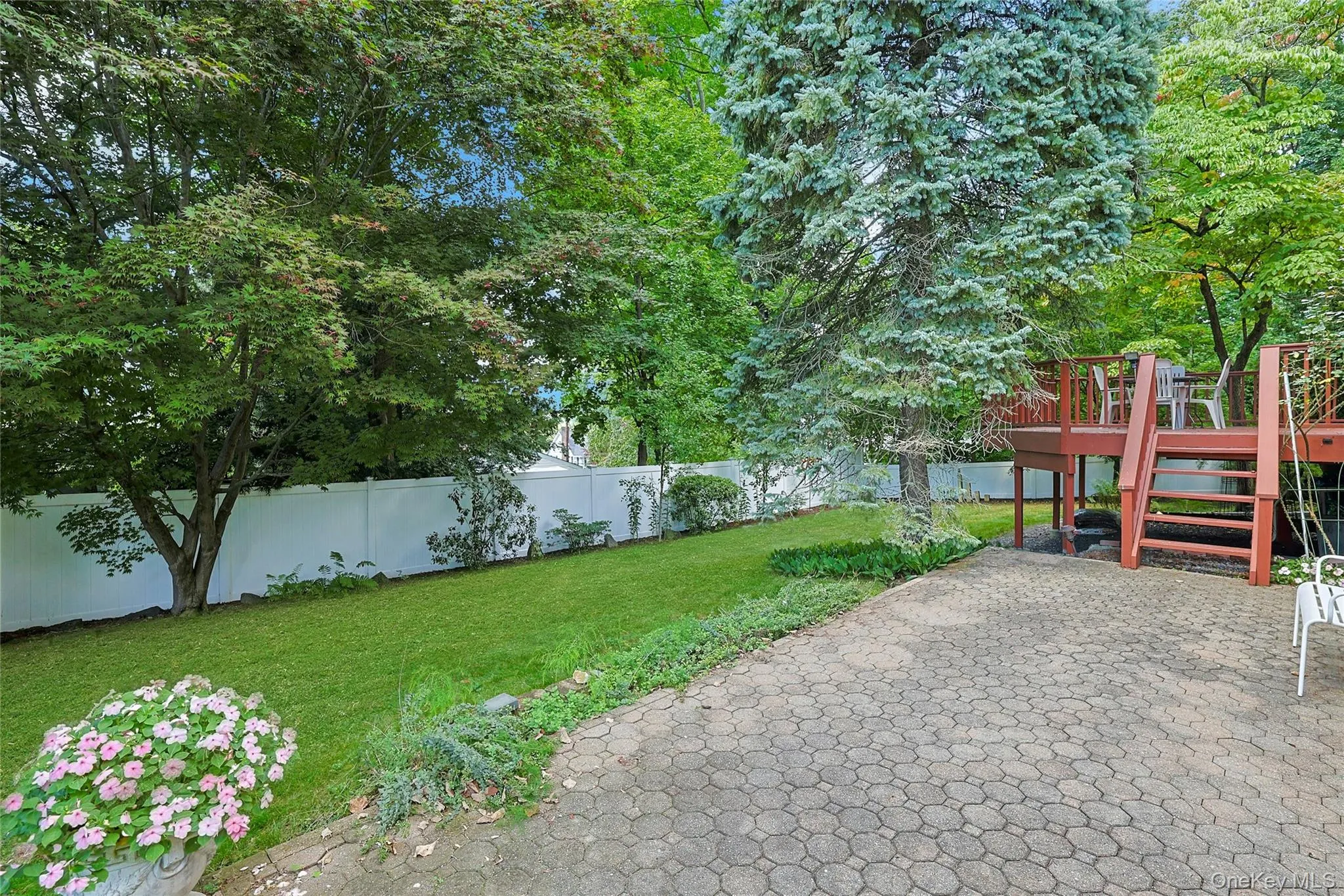 Another view of the expansive backyard featuring a manicured lawn, established trees, a paver patio, steps up to the wood deck, and a white privacy fence. Another view of the expansive backyard featuring a manicured lawn, established trees, a paver patio, steps up to the wood deck, and a white privacy fence.