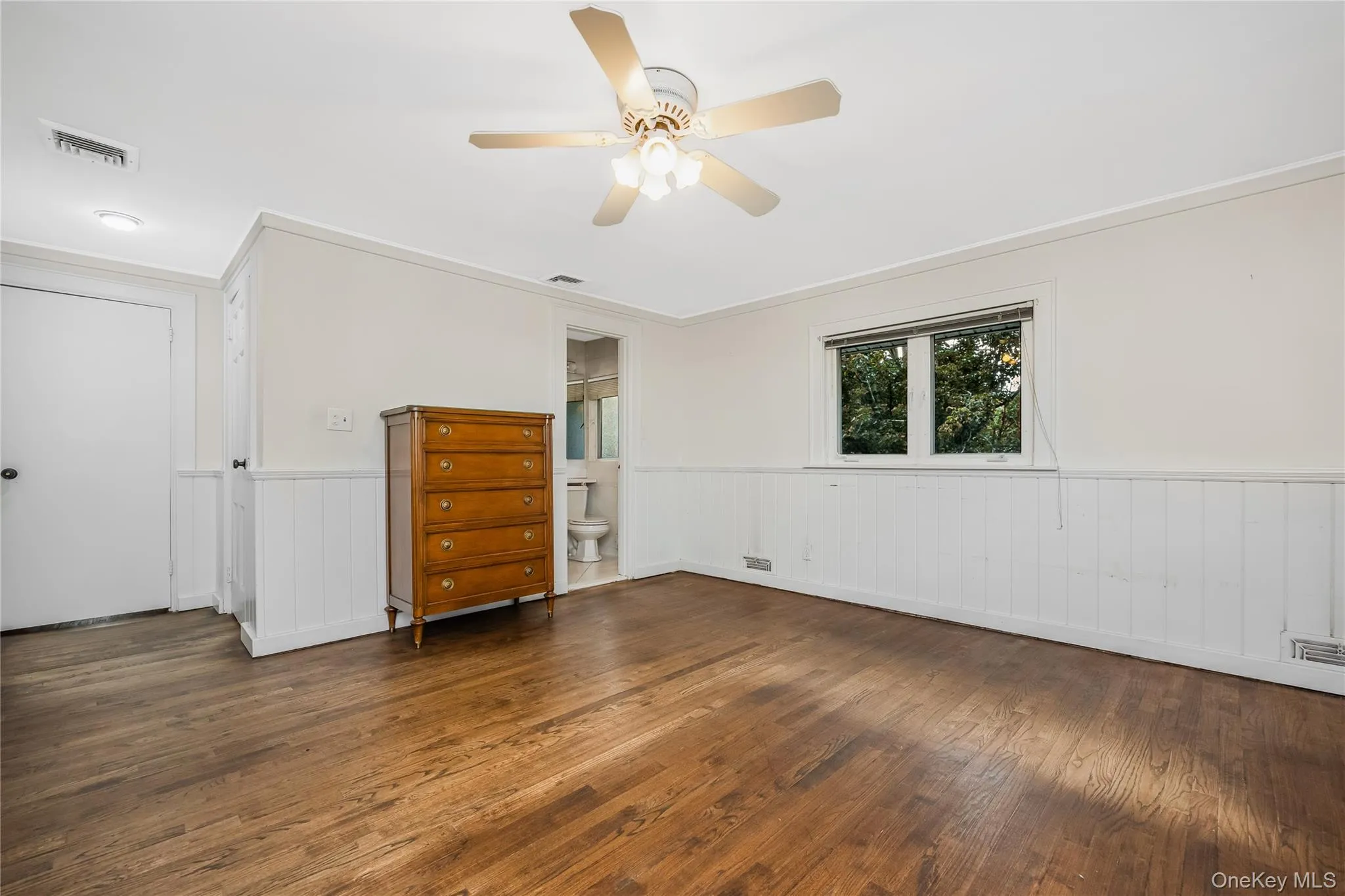 Wood-finish flooring, white paneled wainscoting, and a ceiling fan with integrated lighting and access to the primary bath Wood-finish flooring, white paneled wainscoting, and a ceiling fan with integrated lighting and access to the primary bath
