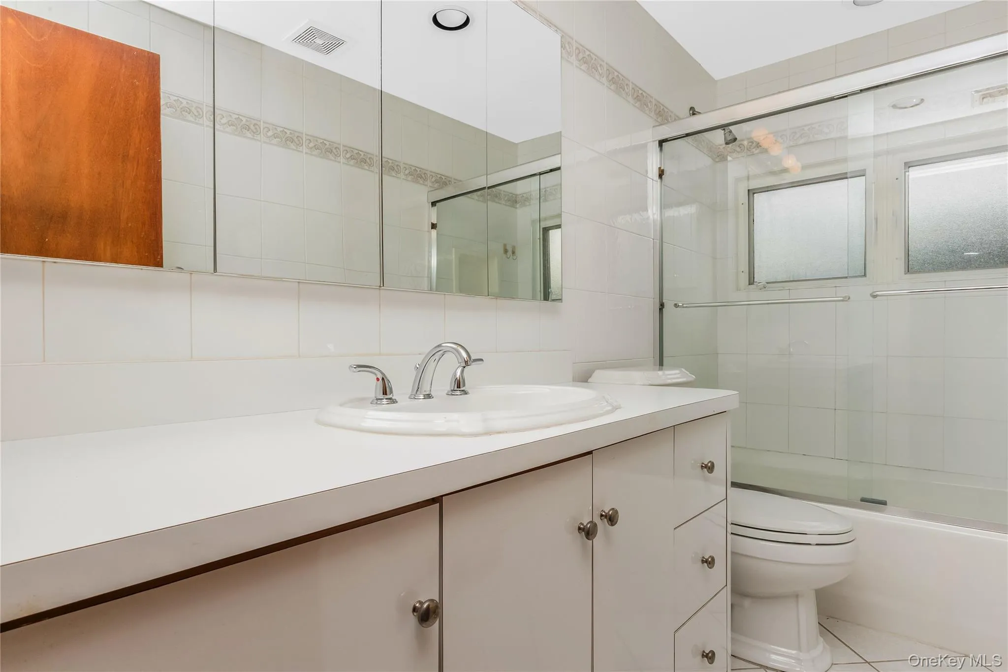 Hall Bath. White vanity integrated sink, chrome faucet, mirrored medicine cabinet, built-in bathtub with glass enclosure, and white tiled walls Hall Bath. White vanity integrated sink, chrome faucet, mirrored medicine cabinet, built-in bathtub with glass enclosure, and white tiled walls