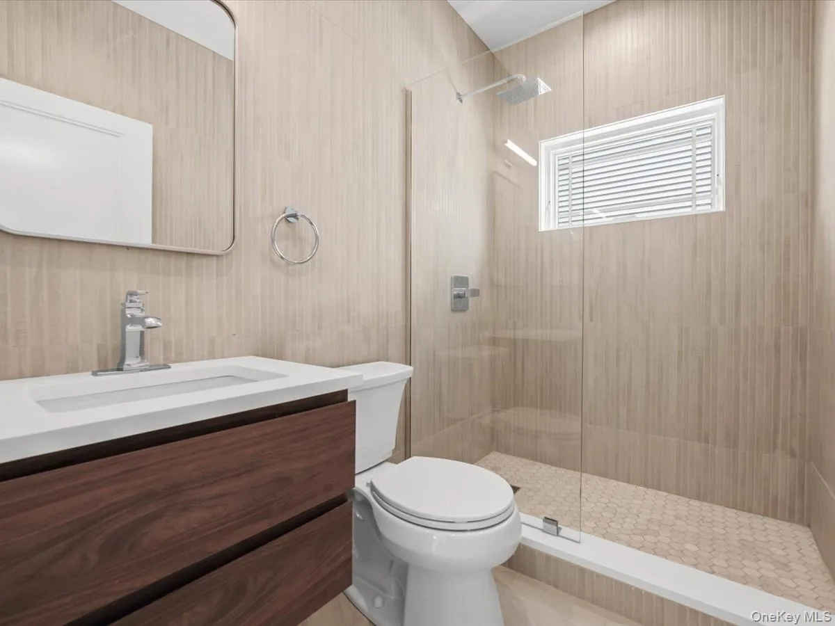 Floating wood-finish vanity with an integrated rectangular sink, clear glass shower enclosure, rainfall showerhead, and light-toned vertical tile Floating wood-finish vanity with an integrated rectangular sink, clear glass shower enclosure, rainfall showerhead, and light-toned vertical tile