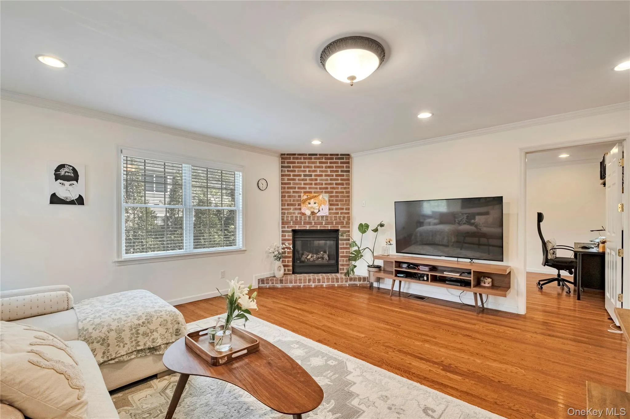 Living room featuring wood finished floors, crown molding, a brick fireplace, recessed lighting, and a desk Living room featuring wood finished floors, crown molding, a brick fireplace, recessed lighting, and a desk