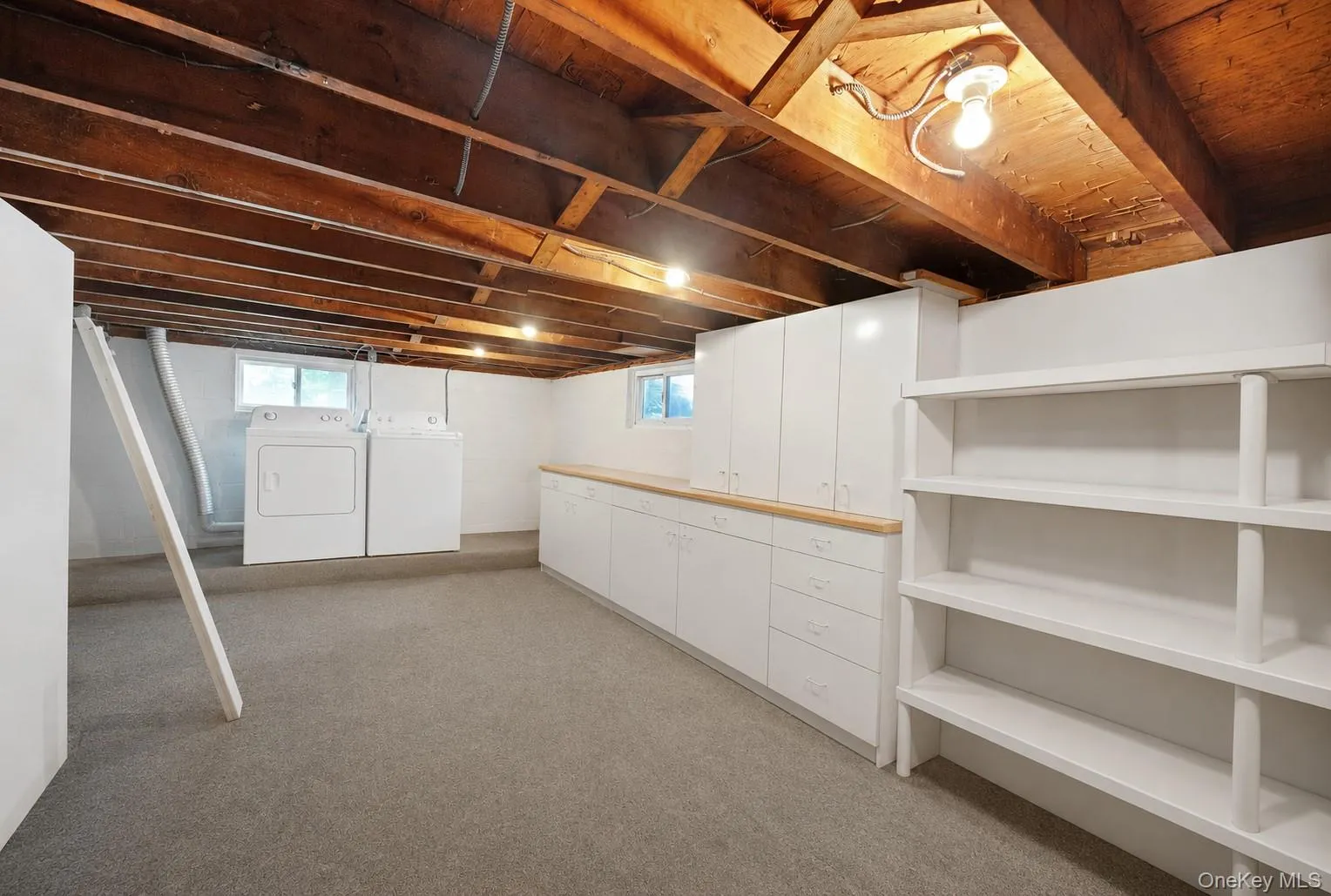 Lower level utility space featuring exposed wood beam ceiling, plush carpet flooring, white cabinetry with wood-finish countertops, and built-in shelving Lower level utility space featuring exposed wood beam ceiling, plush carpet flooring, white cabinetry with wood-finish countertops, and built-in shelving