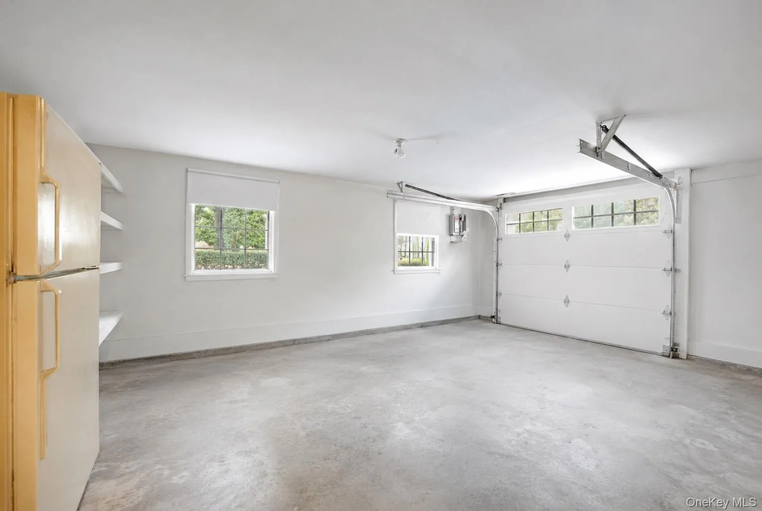 Spacious garage featuring a concrete floor, two windows with roller shades, and a white paneled garage door with upper window inserts Spacious garage featuring a concrete floor, two windows with roller shades, and a white paneled garage door with upper window inserts