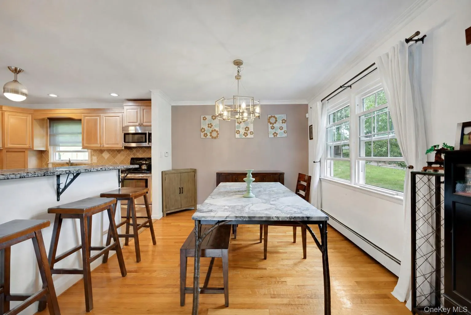 Open-concept dining area featuring wood-finish flooring, a contemporary chandelier, two large windows, a breakfast bar with stone-finish countertop, and light wood-finish cabinetry in the adjacent kitchen Open-concept dining area featuring wood-finish flooring, a contemporary chandelier, two large windows, a breakfast bar with stone-finish countertop, and light wood-finish cabinetry in the adjacent kitchen