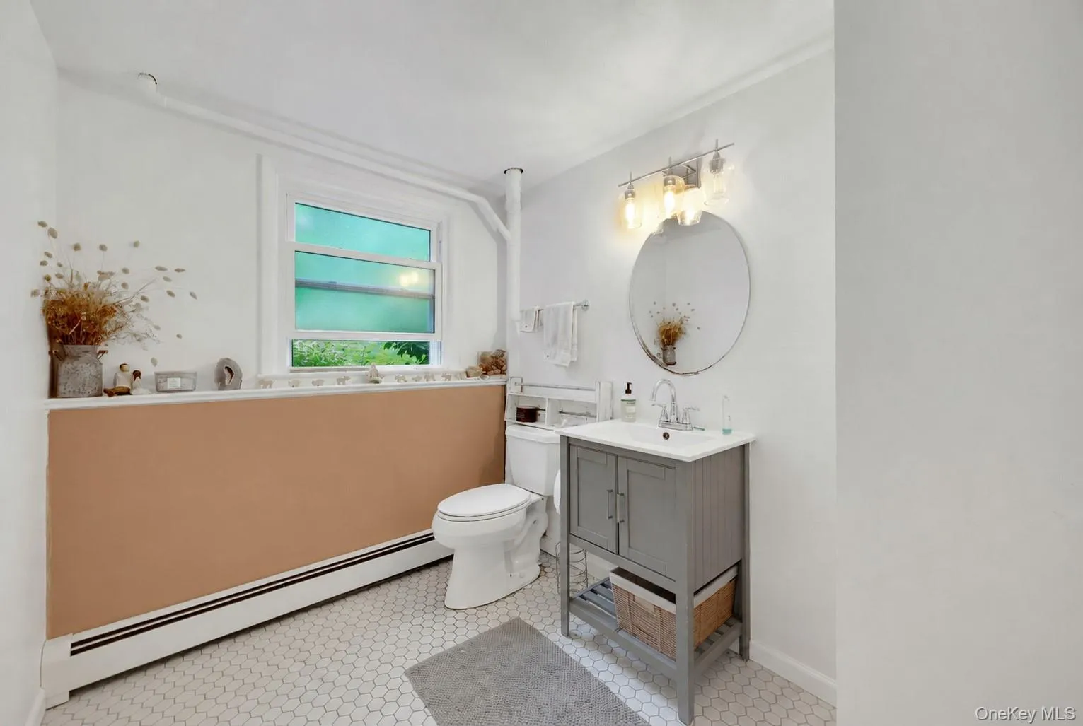 Bathroom featuring white hexagonal floor tiles, a window with white trim, a gray vanity with a white countertop and integrated sink, a round mirror, and a three-light vanity fixture Bathroom featuring white hexagonal floor tiles, a window with white trim, a gray vanity with a white countertop and integrated sink, a round mirror, and a three-light vanity fixture