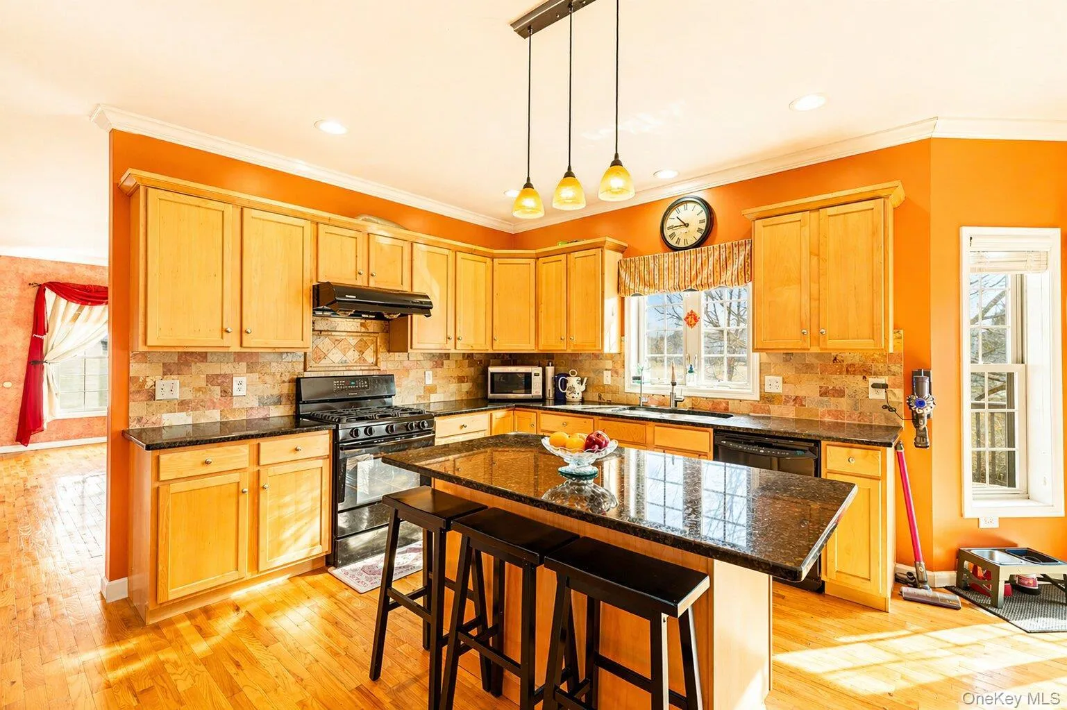 Open-concept kitchen featuring wood-finish flooring, a central island with countertop seating, light wood cabinetry, stone backsplash, and black appliances Open-concept kitchen featuring wood-finish flooring, a central island with countertop seating, light wood cabinetry, stone backsplash, and black appliances