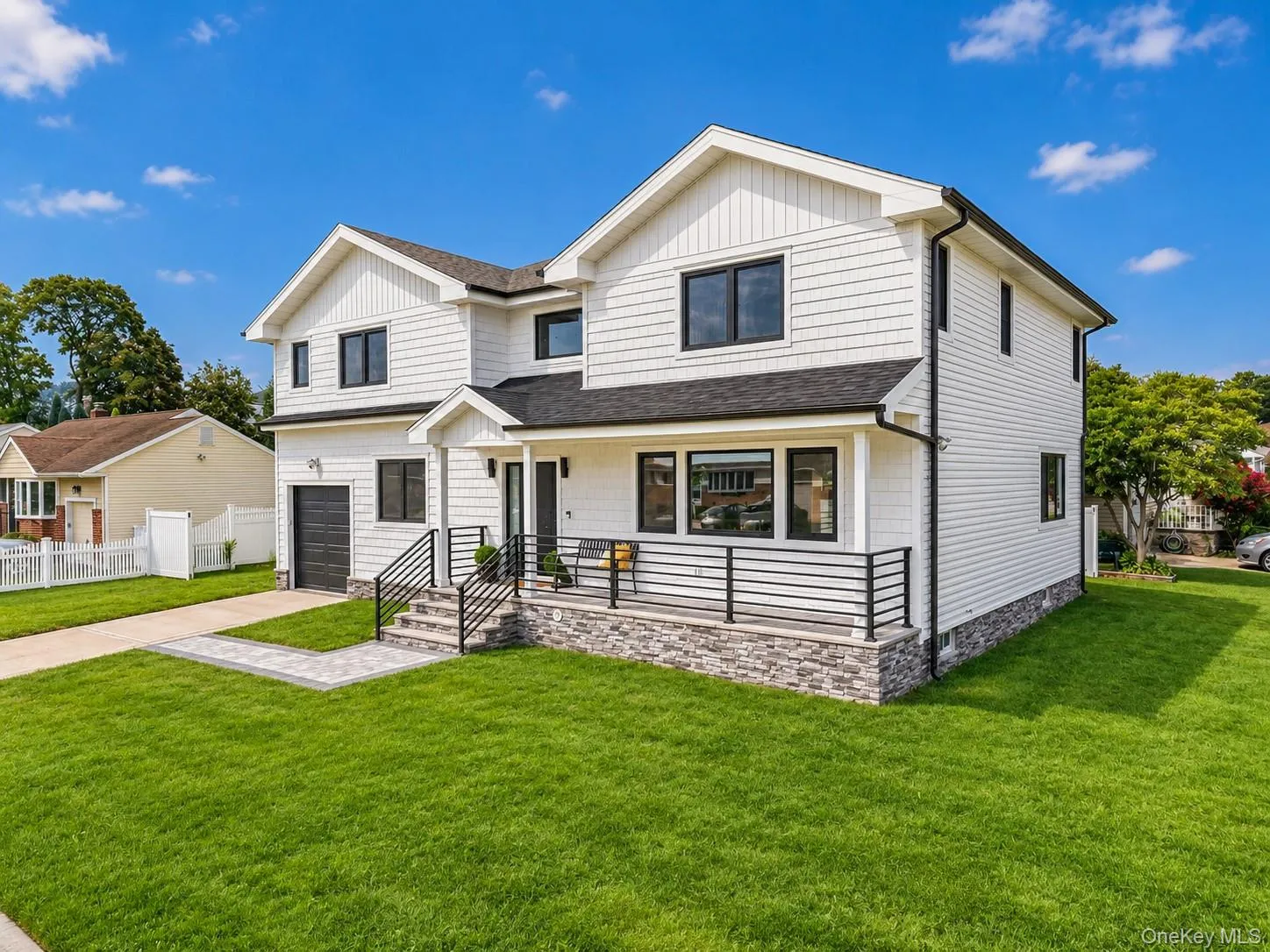 White exterior with dark trim windows, covered porch with stone detailing, black metal railings, attached garage, and a manicured lawn White exterior with dark trim windows, covered porch with stone detailing, black metal railings, attached garage, and a manicured lawn