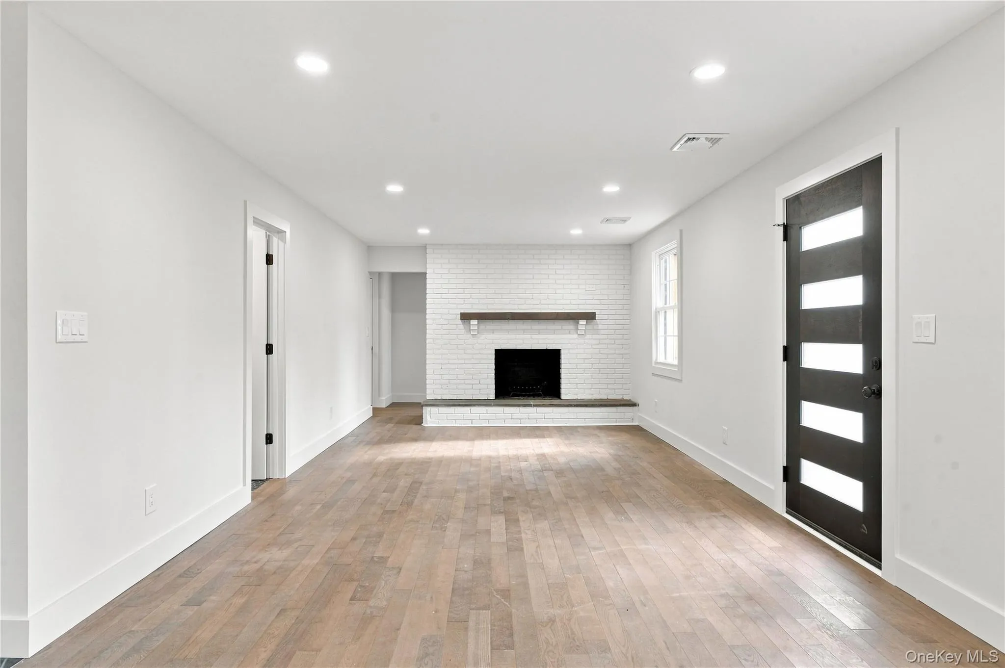 Expansive room with wood-finish flooring, a white brick fireplace with a wood mantle, recessed lighting, and a modern entry door with frosted glass panels Expansive room with wood-finish flooring, a white brick fireplace with a wood mantle, recessed lighting, and a modern entry door with frosted glass panels