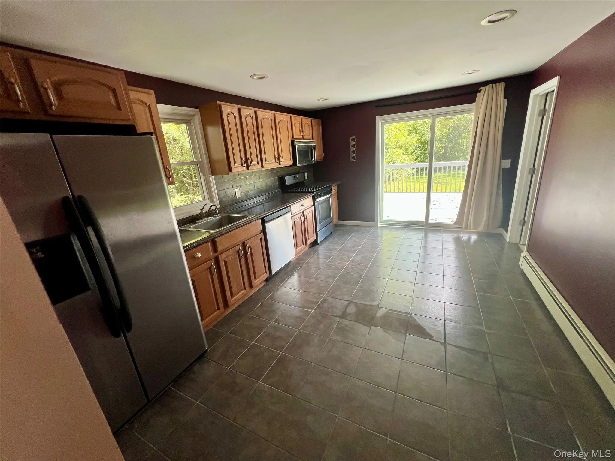 Kitchen featuring wood cabinetry, stainless steel appliances, dark tile flooring, and a sliding glass door to an outdoor deck Kitchen featuring wood cabinetry, stainless steel appliances, dark tile flooring, and a sliding glass door to an outdoor deck