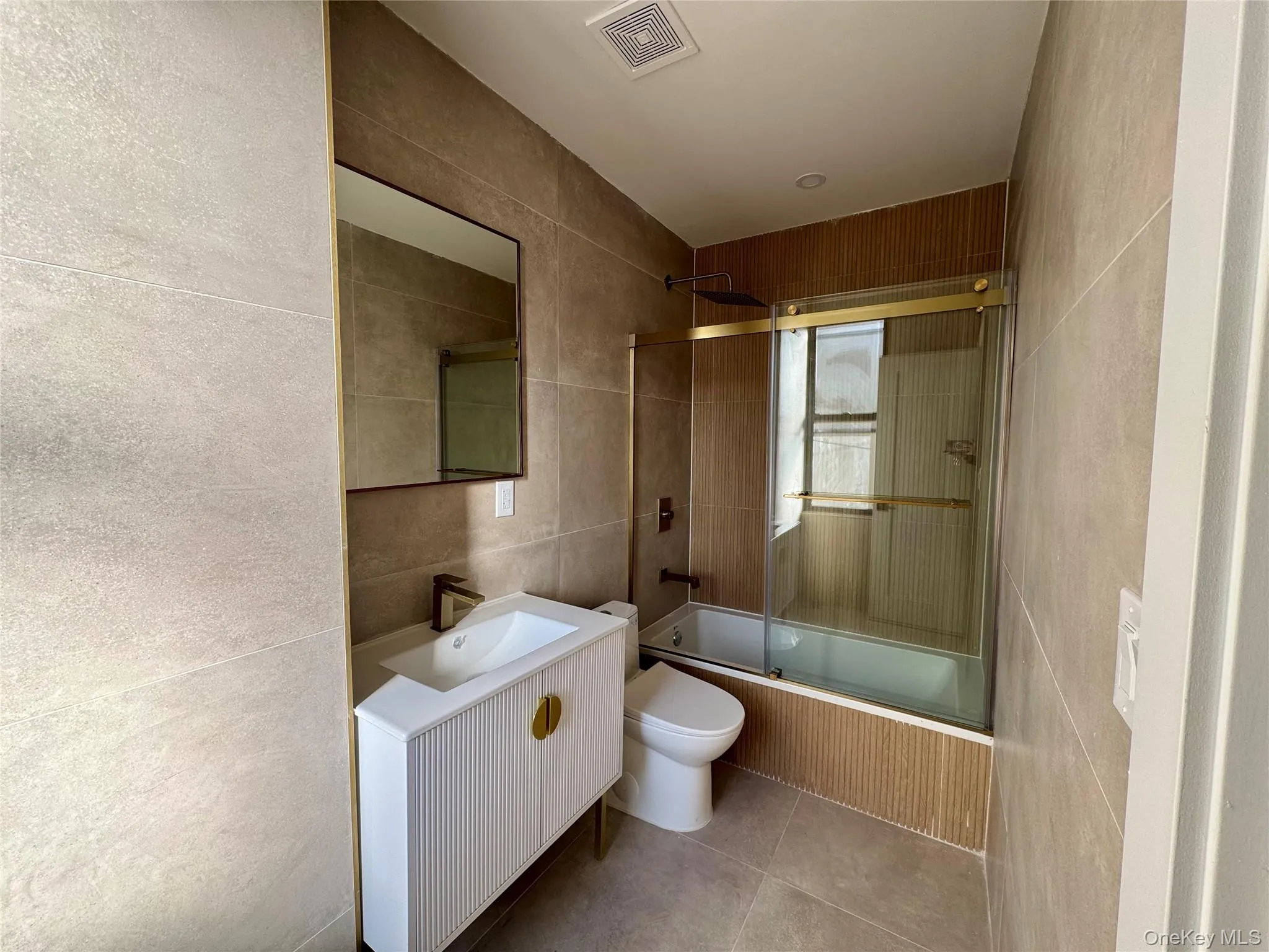 Full bathroom with tile patterned floors, bath / shower combo with glass door, toilet, vanity, and tile walls Full bathroom with tile patterned floors, bath / shower combo with glass door, toilet, vanity, and tile walls