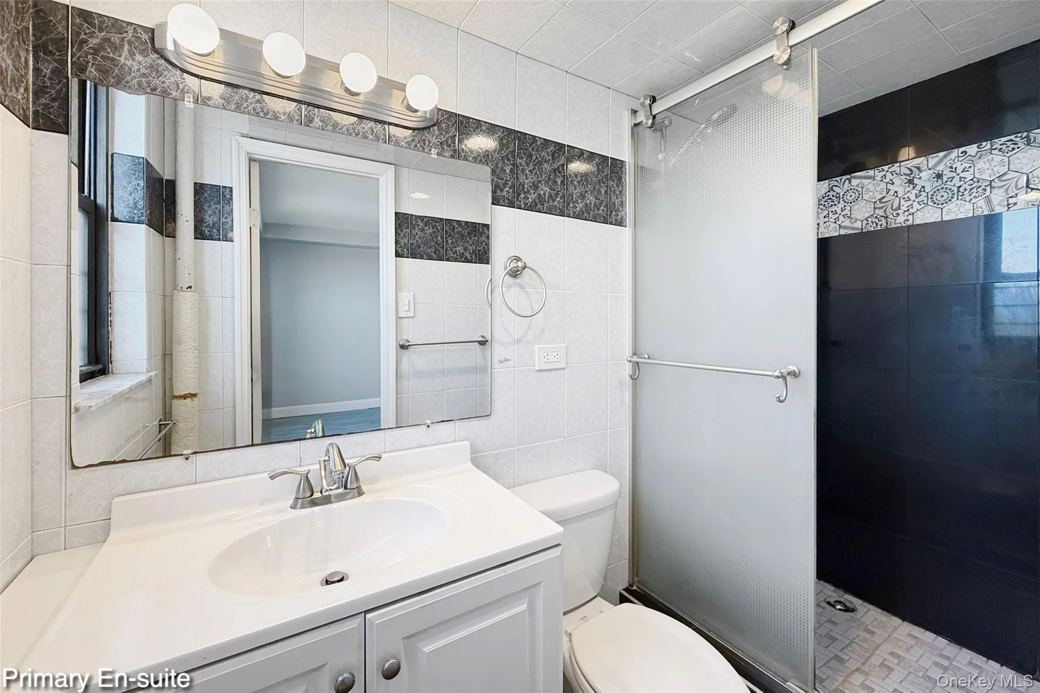 White vanity with an integrated sink and chrome faucet, large wall mirror with strip lighting, tiled walls, and a glass-enclosed shower with decorative tile accents White vanity with an integrated sink and chrome faucet, large wall mirror with strip lighting, tiled walls, and a glass-enclosed shower with decorative tile accents