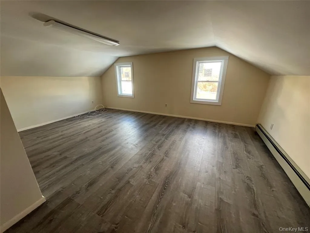 Spacious room featuring wood-finish flooring, an angled ceiling, two windows, and a fluorescent ceiling light fixture Spacious room featuring wood-finish flooring, an angled ceiling, two windows, and a fluorescent ceiling light fixture