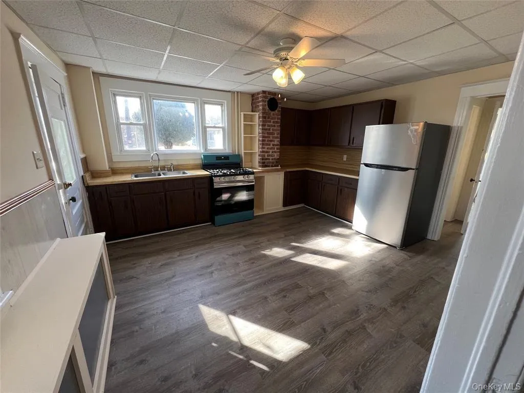 Wood-finish flooring, dark wood cabinetry, a stainless steel refrigerator, and an exposed brick accent wall Wood-finish flooring, dark wood cabinetry, a stainless steel refrigerator, and an exposed brick accent wall