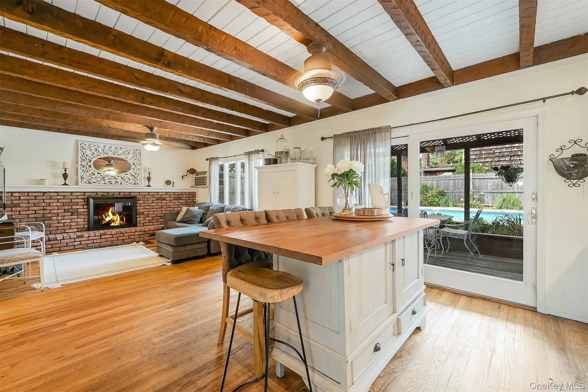 Hardwood flooring, exposed wood beam ceiling, brick fireplace, built-in kitchen island with wood countertop, and sliding glass doors to outdoor area with a pool Hardwood flooring, exposed wood beam ceiling, brick fireplace, built-in kitchen island with wood countertop, and sliding glass doors to outdoor area with a pool