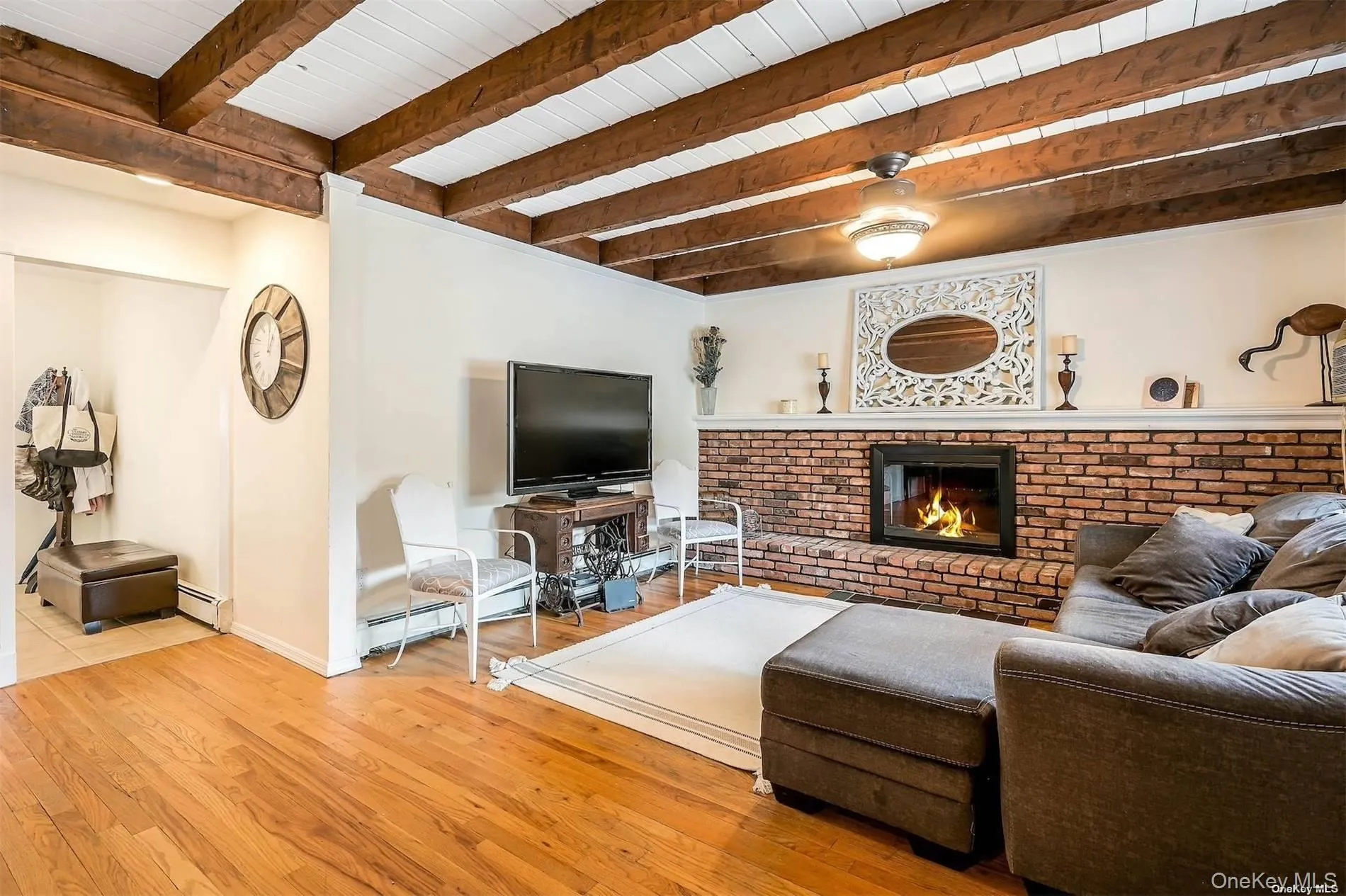 Exposed wood beam ceiling with white panels, brick-clad fireplace with a hearth, wood-finish flooring, and ceiling fan with integrated lighting Exposed wood beam ceiling with white panels, brick-clad fireplace with a hearth, wood-finish flooring, and ceiling fan with integrated lighting