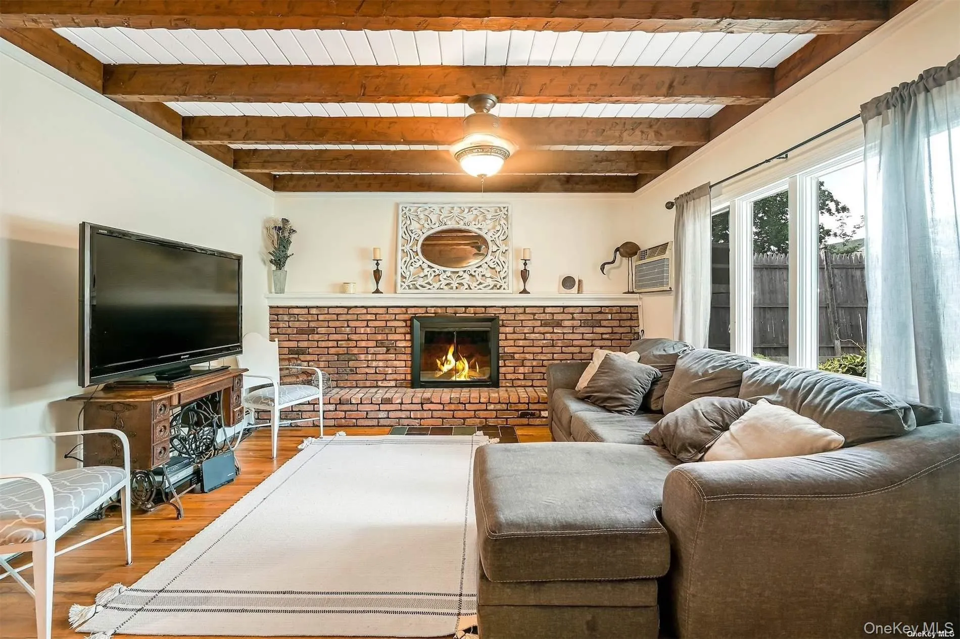 Exposed wood beam ceiling with white panels, brick-surround fireplace, wood-finish flooring, and a large window providing outdoor illumination Exposed wood beam ceiling with white panels, brick-surround fireplace, wood-finish flooring, and a large window providing outdoor illumination