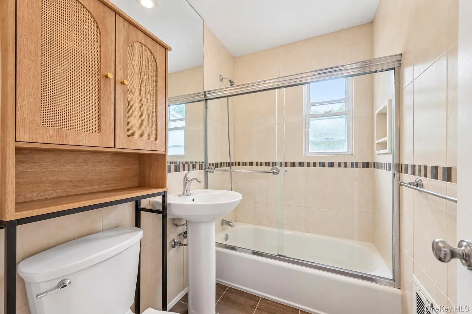 Pedestal sink with chrome faucet, shower-over-tub with sliding glass enclosure, neutral tile surround, and wood-finish overhead cabinetry Pedestal sink with chrome faucet, shower-over-tub with sliding glass enclosure, neutral tile surround, and wood-finish overhead cabinetry