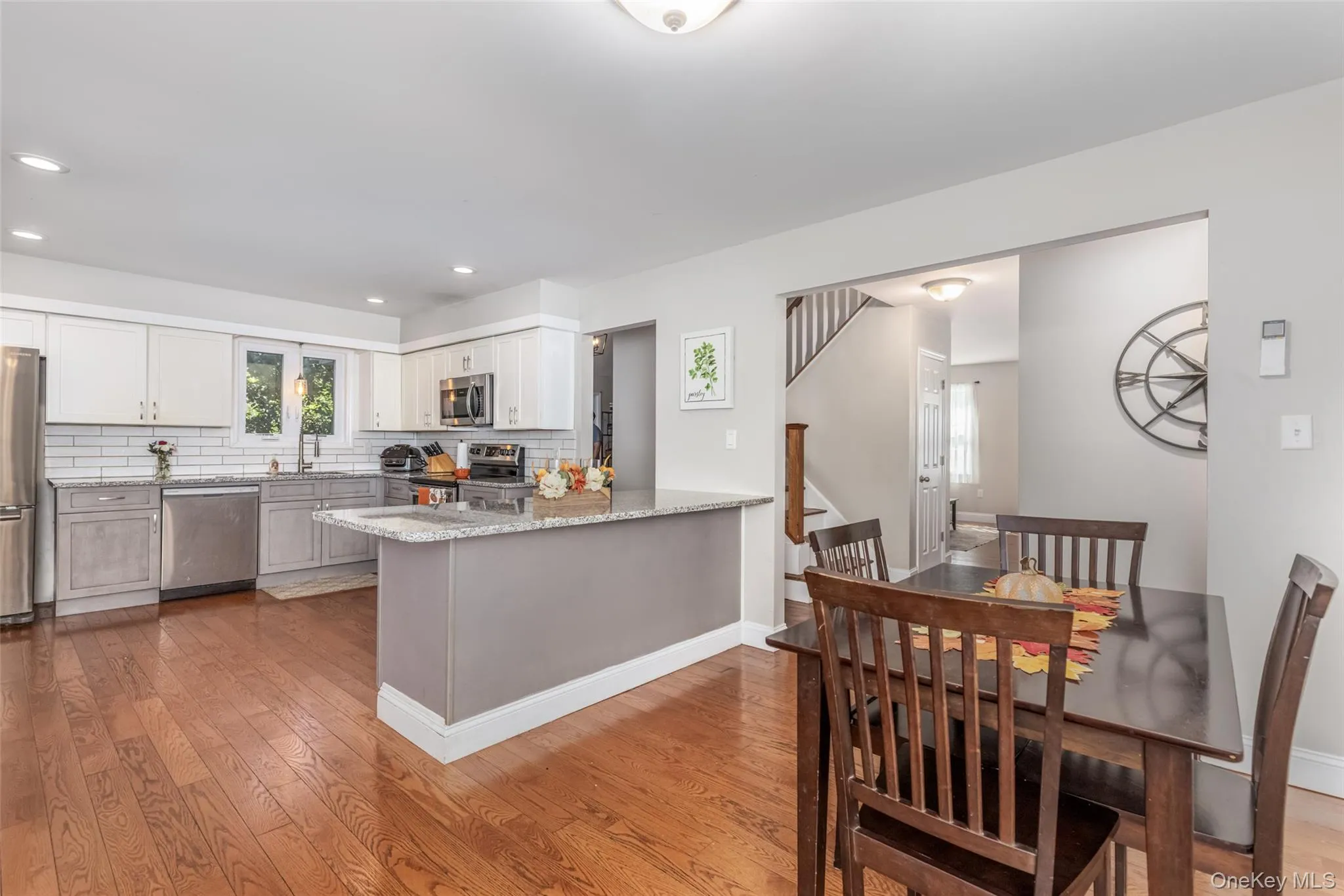 Wood-finish flooring throughout, kitchen featuring white shaker cabinetry, stainless steel appliances, and a central island with a light-toned countertop Wood-finish flooring throughout, kitchen featuring white shaker cabinetry, stainless steel appliances, and a central island with a light-toned countertop