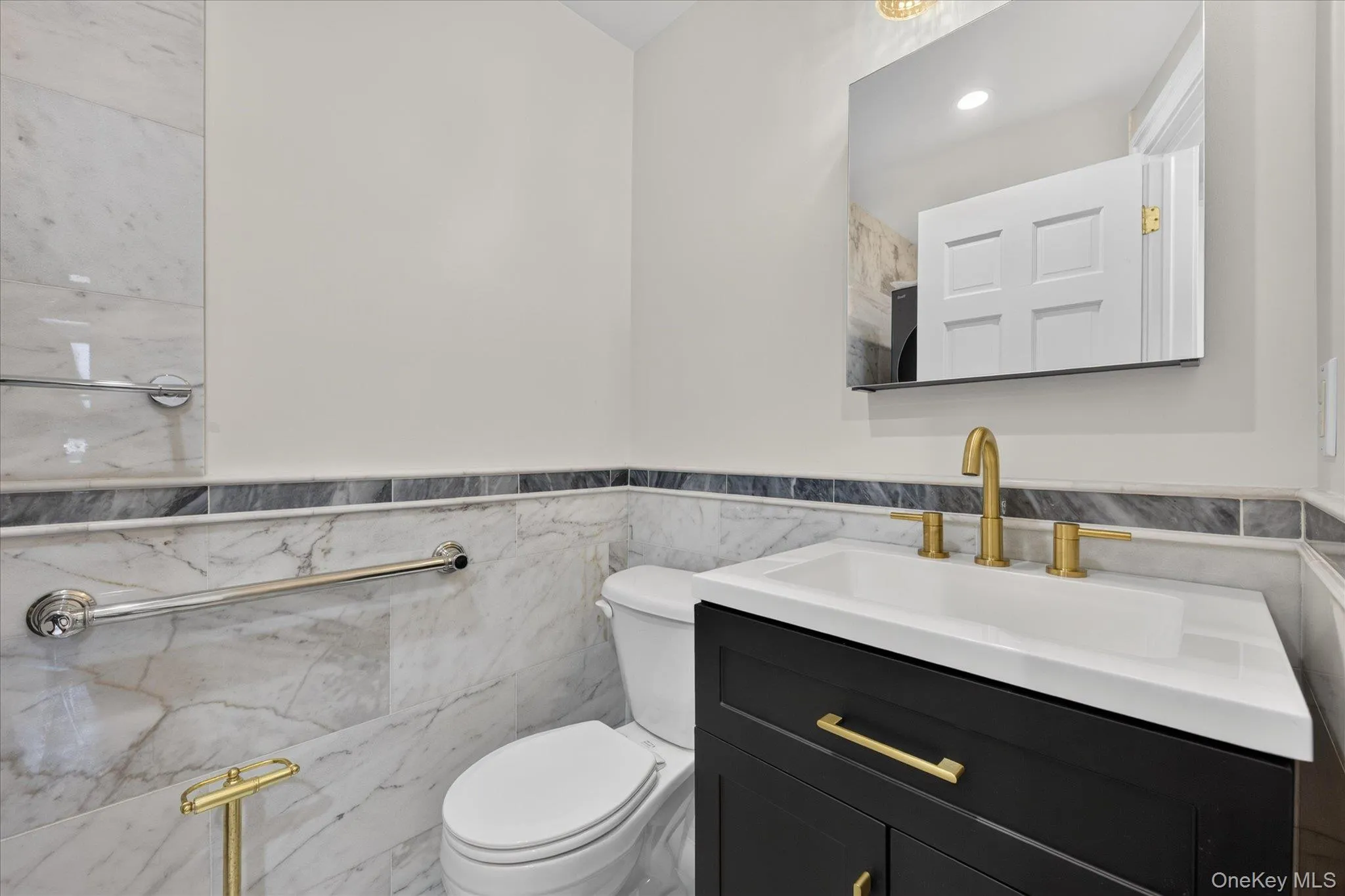 Half bathroom on first floor has a vanity with a white integrated sink and gold-finish hardware, gold-finish faucet, wall-mounted mirror, and marble-look wall tiling Half bathroom on first floor has a vanity with a white integrated sink and gold-finish hardware, gold-finish faucet, wall-mounted mirror, and marble-look wall tiling