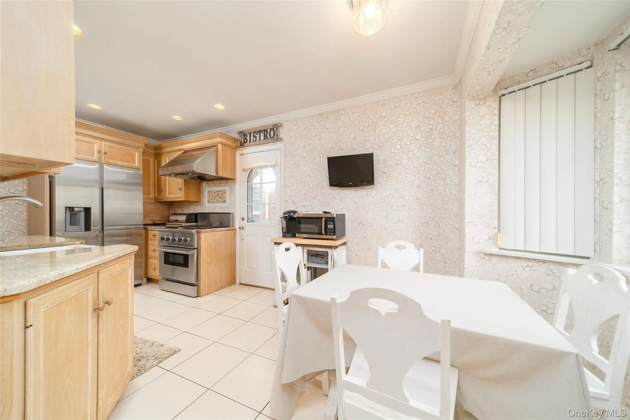 Kitchen featuring stainless steel appliances, light brown cabinets, wall chimney exhaust hood, ornamental molding, and light tile patterned flooring Kitchen featuring stainless steel appliances, light brown cabinets, wall chimney exhaust hood, ornamental molding, and light tile patterned flooring