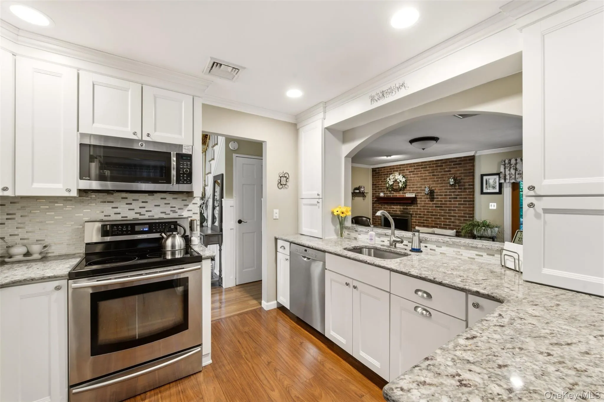 Kitchen featuring white shaker cabinetry, stainless steel appliances, granite countertops, a tile backsplash, and wood-finish flooring Kitchen featuring white shaker cabinetry, stainless steel appliances, granite countertops, a tile backsplash, and wood-finish flooring
