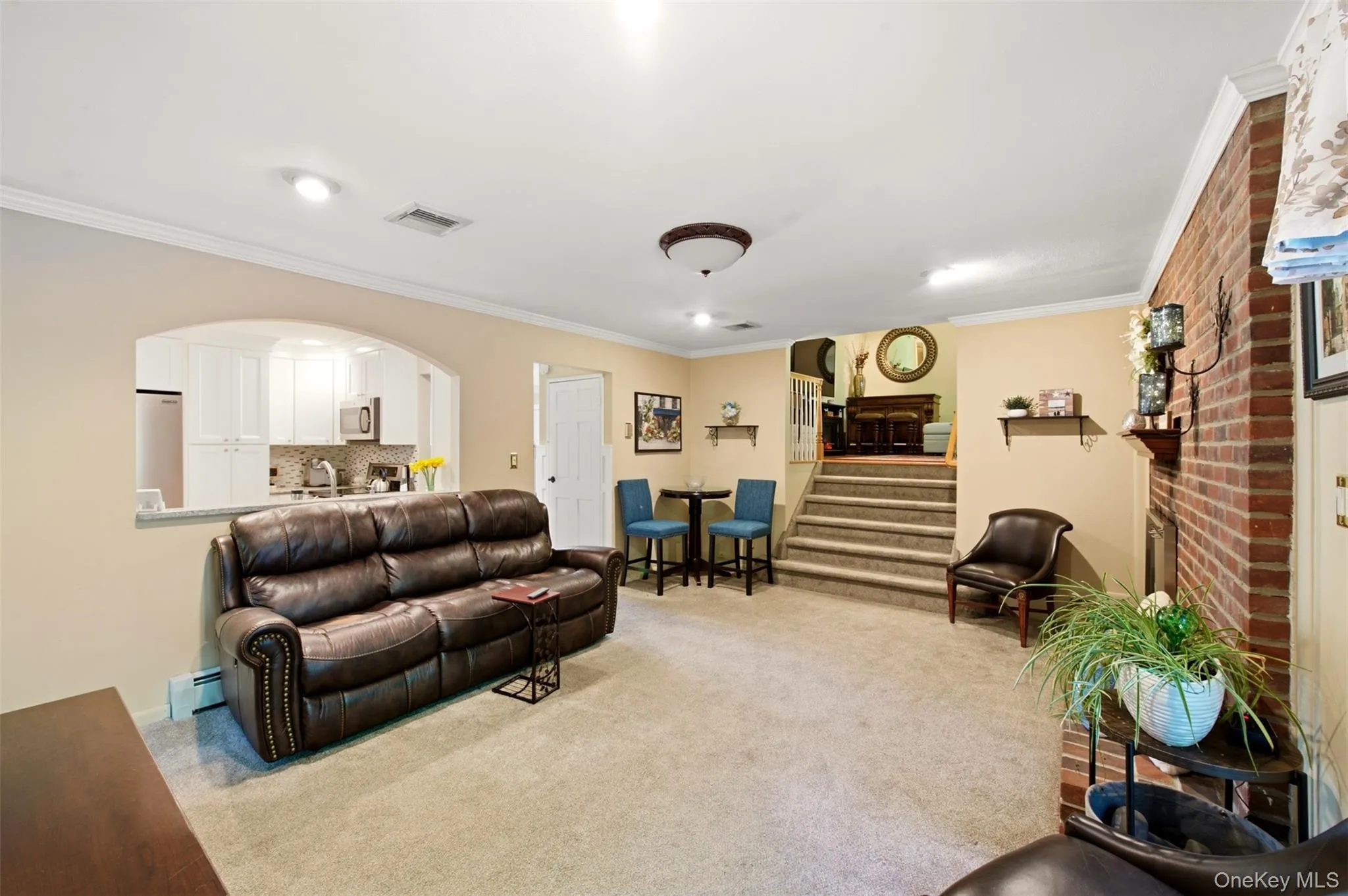 Expansive carpeted living area with a partial brick accent wall and arched pass-through to a kitchen featuring white cabinetry and stainless steel appliances Expansive carpeted living area with a partial brick accent wall and arched pass-through to a kitchen featuring white cabinetry and stainless steel appliances