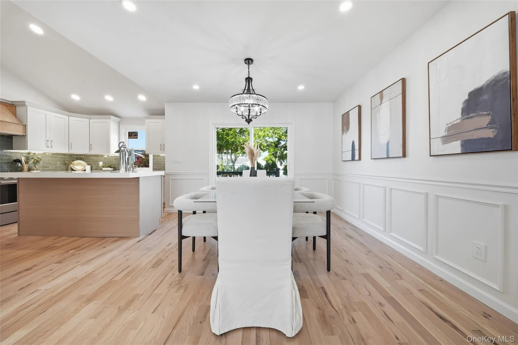 Light wood-finish flooring extending into the kitchen, white wainscoting paneling, and an elegant chandelier Light wood-finish flooring extending into the kitchen, white wainscoting paneling, and an elegant chandelier