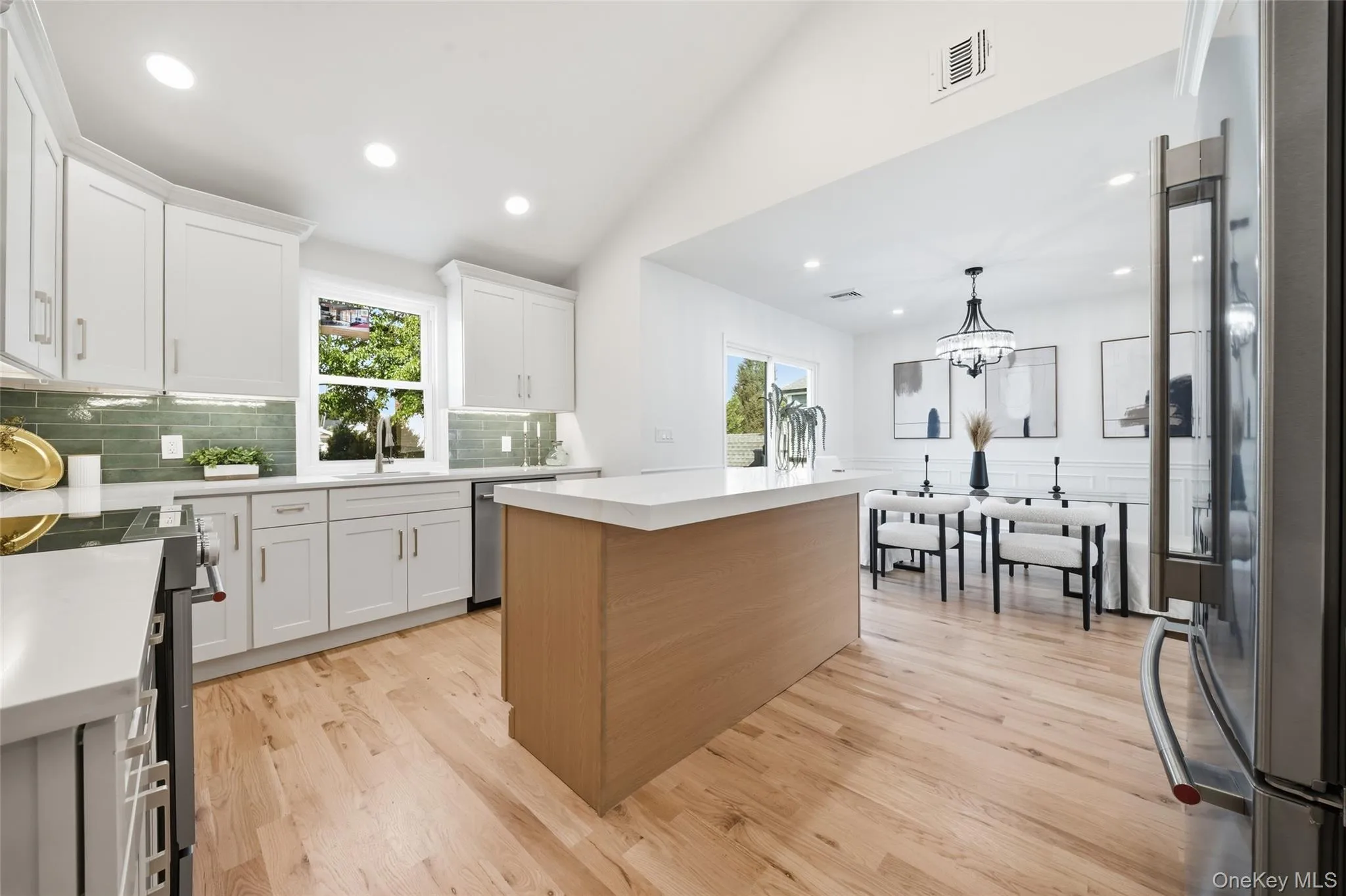 Open-concept kitchen with light wood-finish flooring, white cabinetry, a large kitchen island, and a green subway tile backsplash Open-concept kitchen with light wood-finish flooring, white cabinetry, a large kitchen island, and a green subway tile backsplash