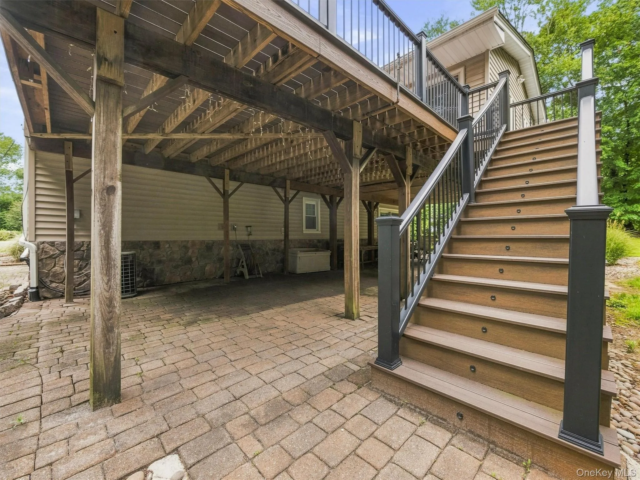 Paver patio beneath a wood-framed deck, featuring stairway with integrated step lighting and dark railing Paver patio beneath a wood-framed deck, featuring stairway with integrated step lighting and dark railing