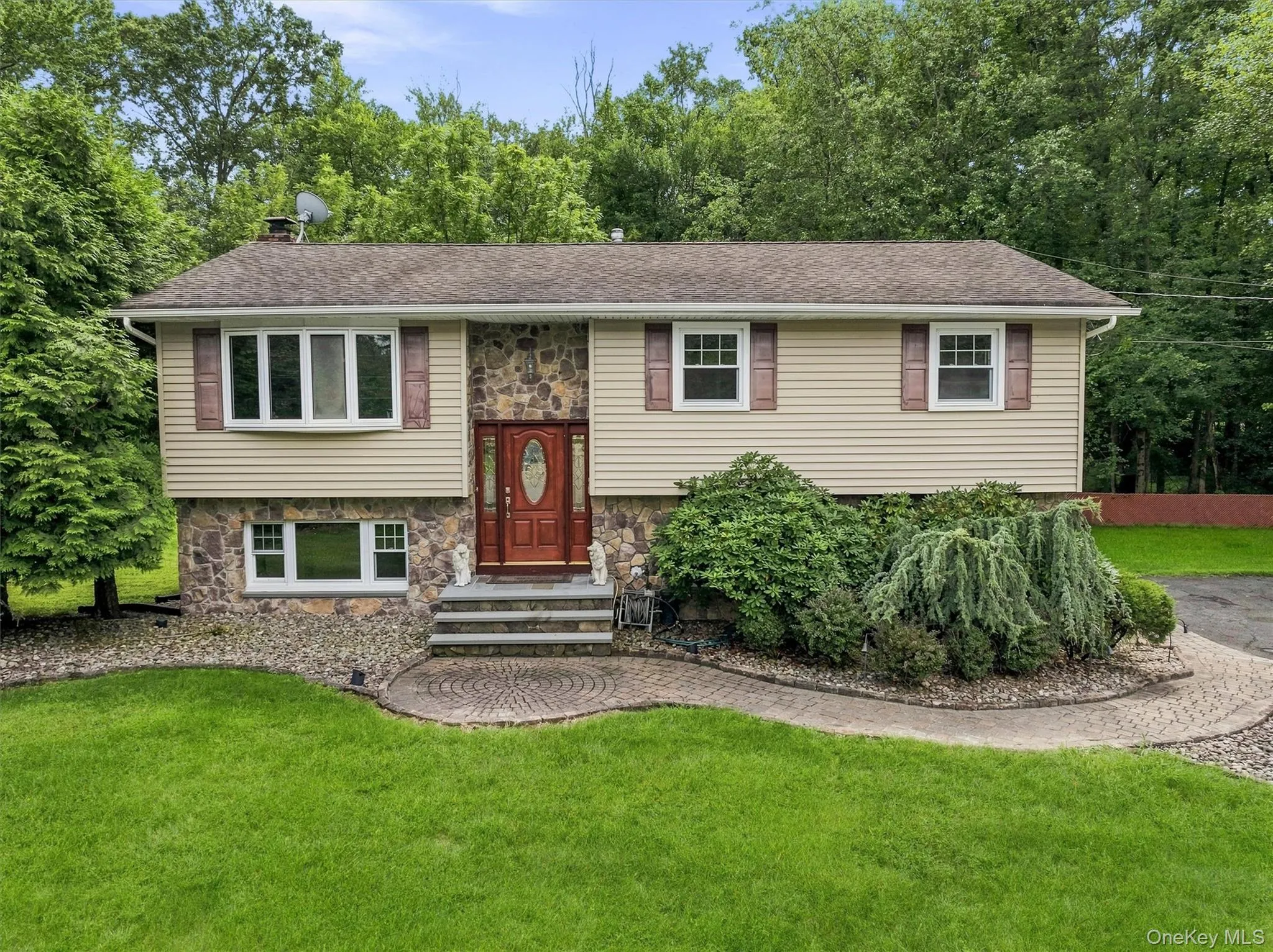 Residence featuring stone and siding facade, bay window, red-toned entry door with oval glass inset, and a paved walkway Residence featuring stone and siding facade, bay window, red-toned entry door with oval glass inset, and a paved walkway