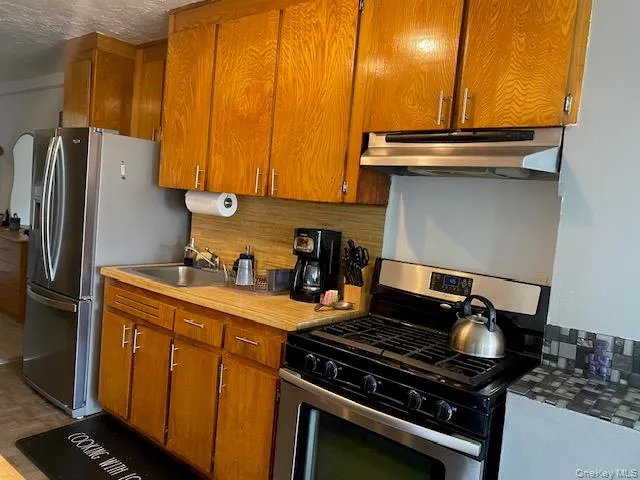 Kitchen featuring tasteful backsplash, a sink, appliances with stainless steel finishes, under cabinet range hood, and brown cabinetry Kitchen featuring tasteful backsplash, a sink, appliances with stainless steel finishes, under cabinet range hood, and brown cabinetry