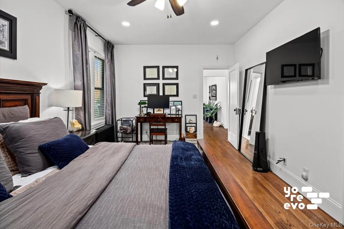 Spacious primary bedroom featuring hardwood flooring, a wood-finish accent wall with dark trim, ceiling fan, wall-mounted flat-screen television, and abundant natural light from large windows with gray drapery Spacious primary bedroom featuring hardwood flooring, a wood-finish accent wall with dark trim, ceiling fan, wall-mounted flat-screen television, and abundant natural light from large windows with gray drapery