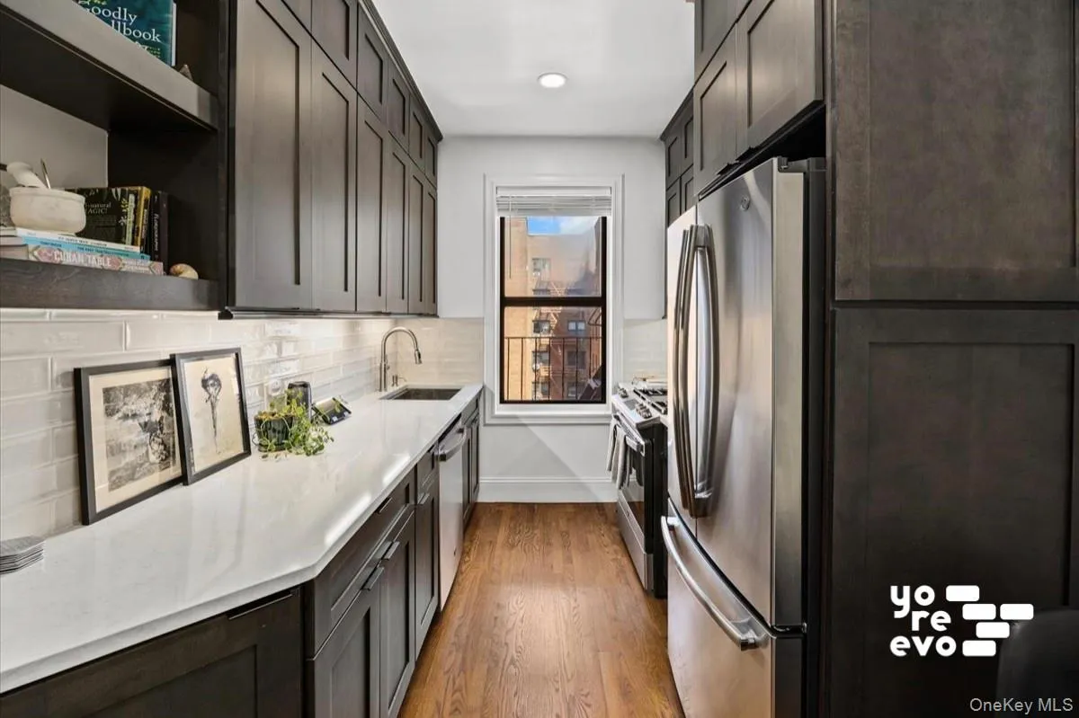 Galley kitchen featuring wood-finish flooring, dark wood-finish cabinetry, light-toned countertops, a subway tile backsplash, and stainless steel appliances Galley kitchen featuring wood-finish flooring, dark wood-finish cabinetry, light-toned countertops, a subway tile backsplash, and stainless steel appliances