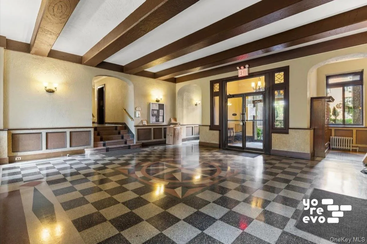 Expansive lobby featuring a checkerboard-patterned floor with a central medallion, exposed ceiling beams, decorative wall sconces, and a multi-pane entry door Expansive lobby featuring a checkerboard-patterned floor with a central medallion, exposed ceiling beams, decorative wall sconces, and a multi-pane entry door